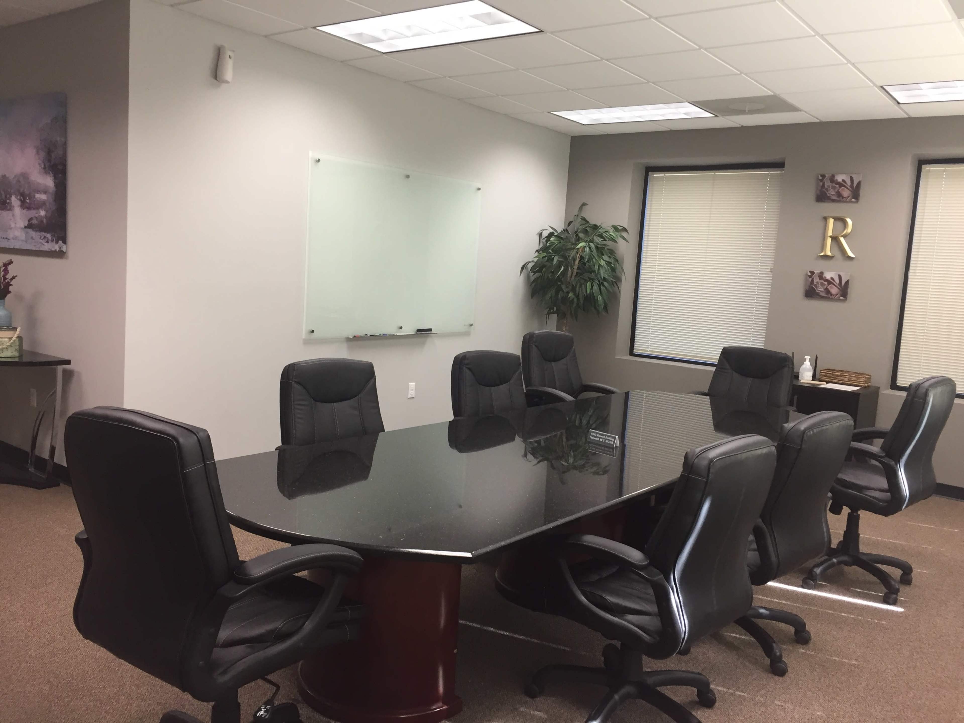 A conference room features a large black table surrounded by eight black leather chairs, with a whiteboard and potted plant in the background.