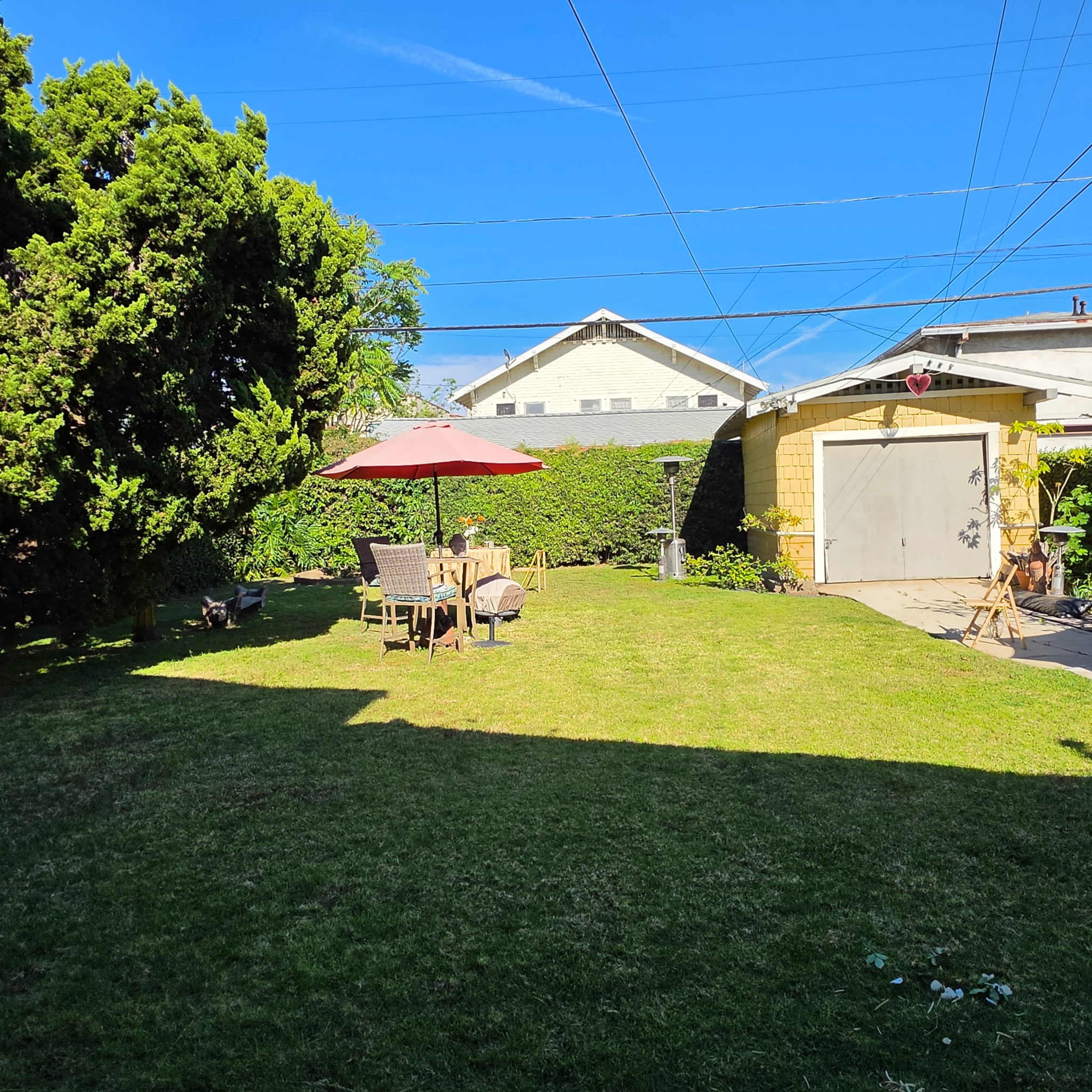 The scene shows a well-maintained backyard with a patio set under an umbrella, surrounded by grassy areas and adjacent buildings.