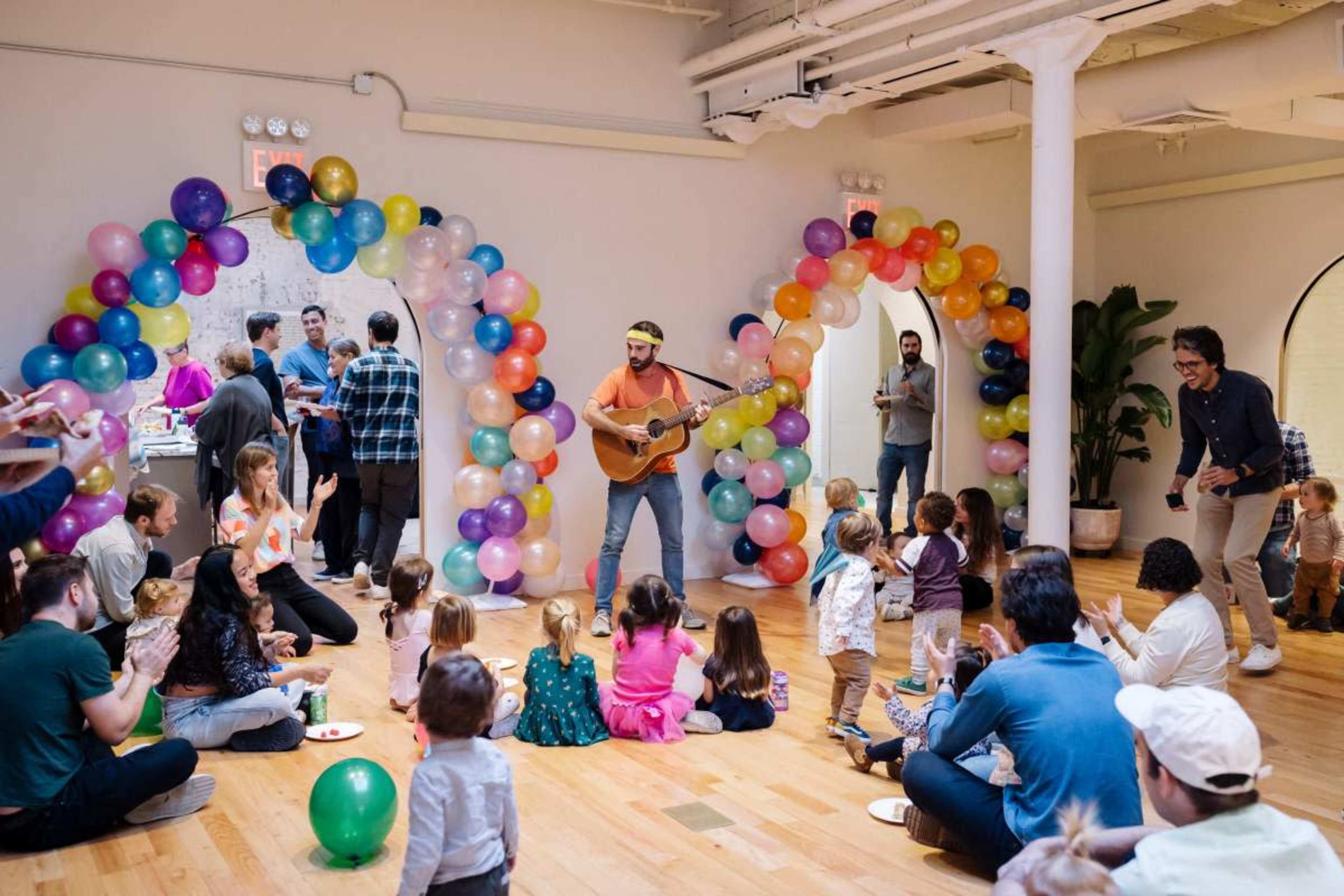 A musician plays guitar for a crowd of children and adults in a room decorated with colorful balloons.