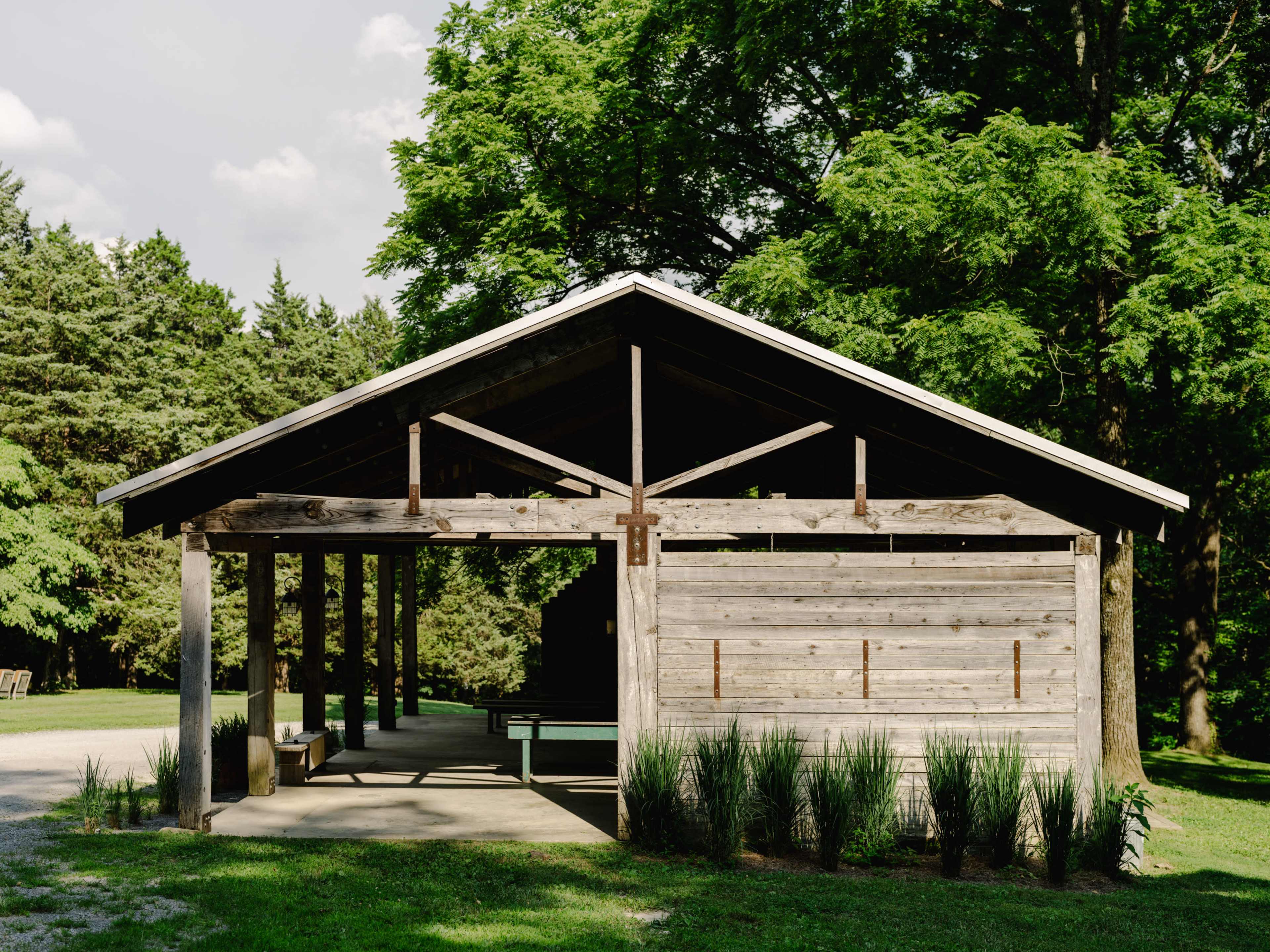 The image shows a rustic wooden pavilion surrounded by green trees and grass.