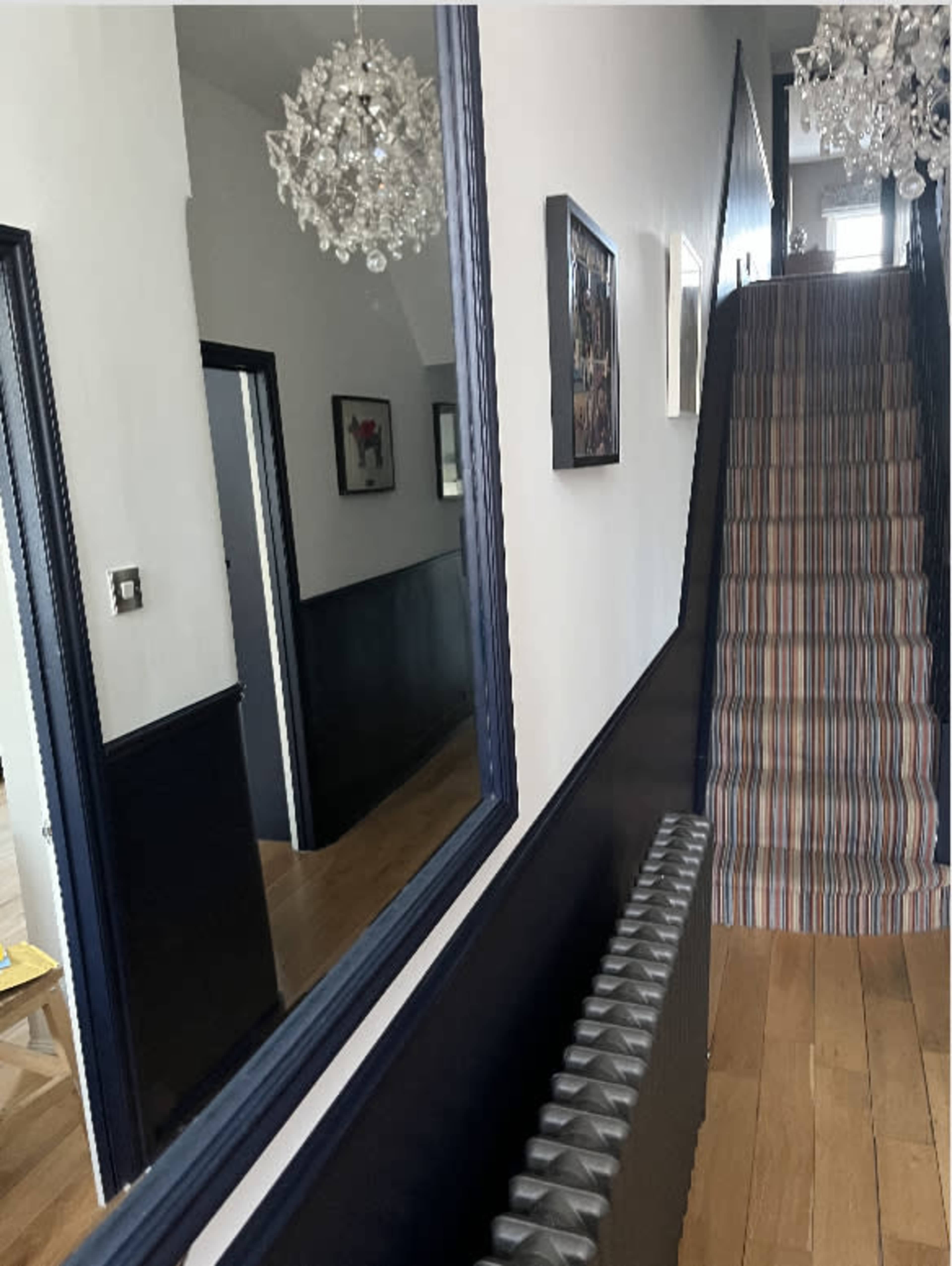 A hallway with a staircase leading upward, featuring a radiator, a mirror on the left wall, and two chandeliers hanging from the ceiling.