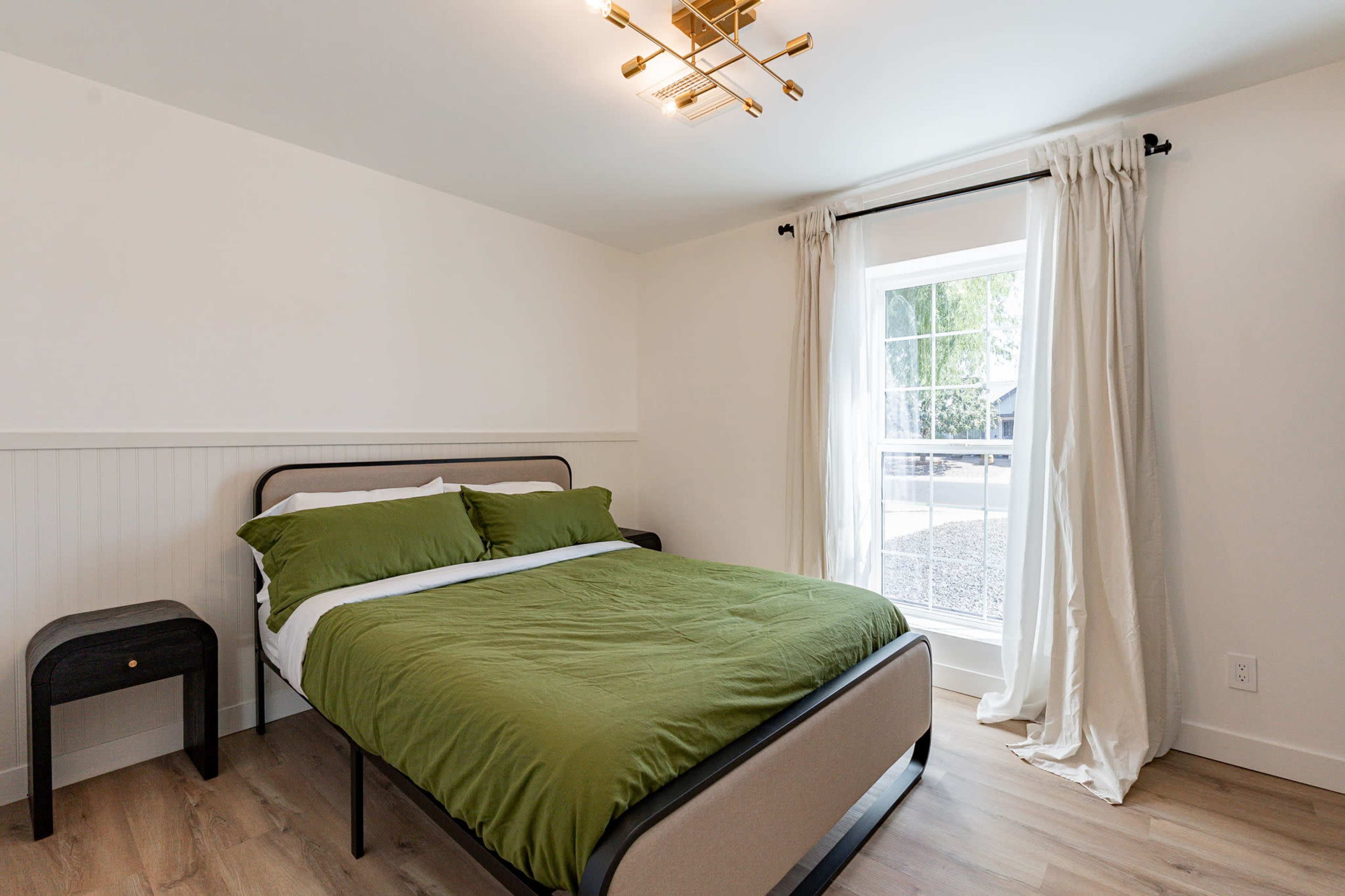 The image shows a minimalist bedroom featuring a bed with a green comforter, a bedside table, and a large window with white curtains.