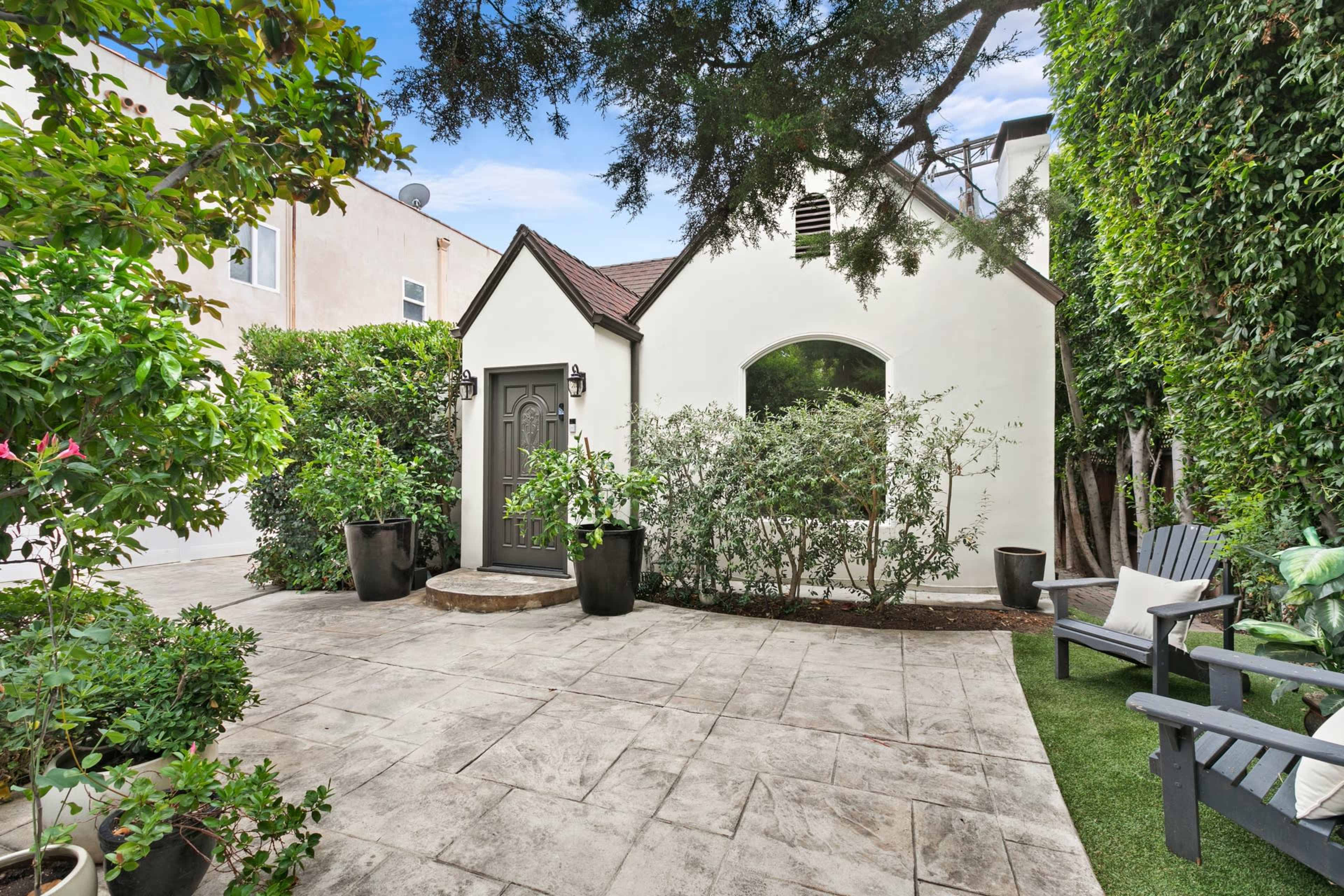 A small, landscaped courtyard features a stone patio surrounded by lush greenery and a charming white cottage with a gray door.