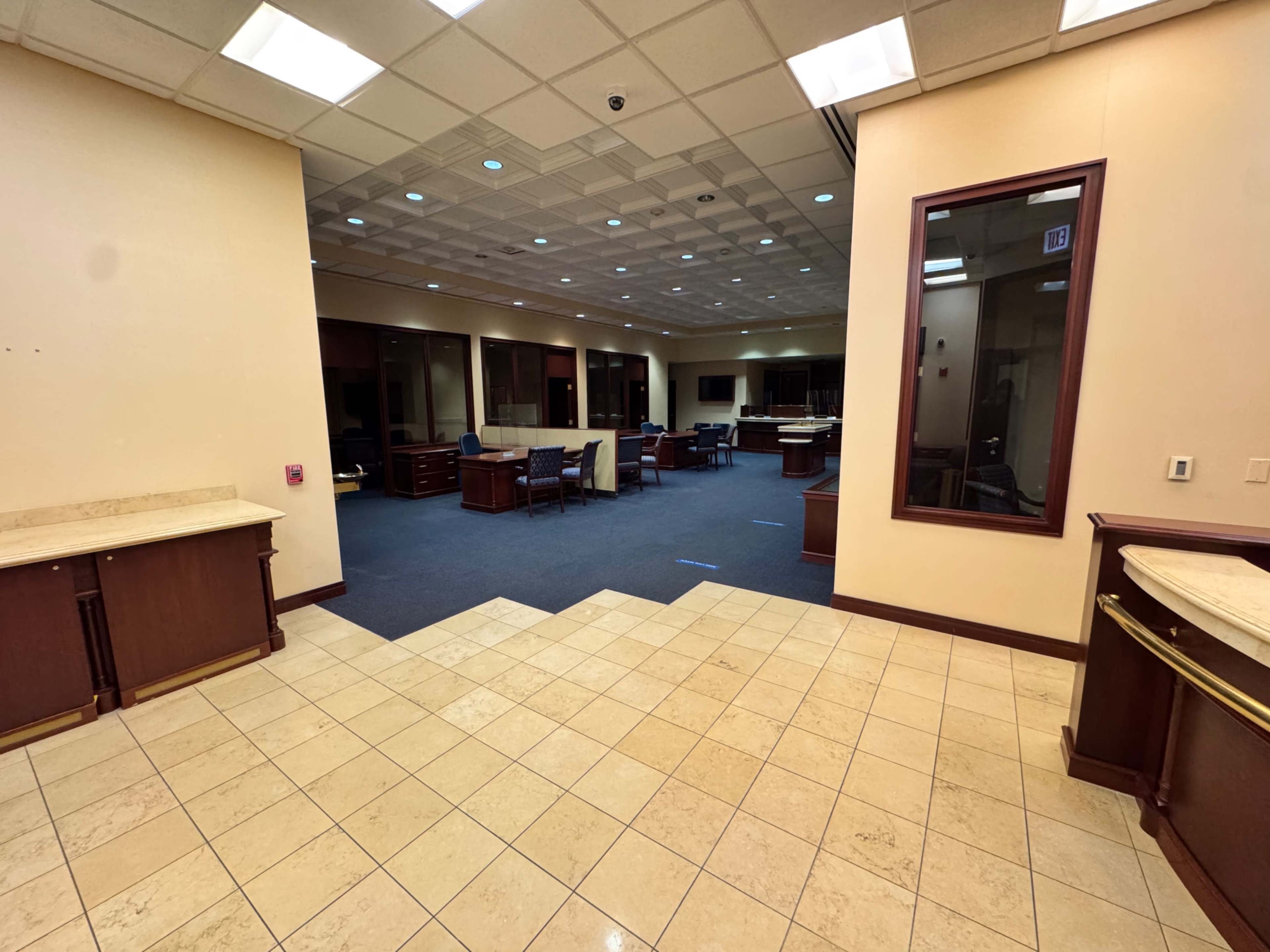 The image shows a large office space with wooden furniture, tiled flooring, and a divided layout into workstations and a reception area.