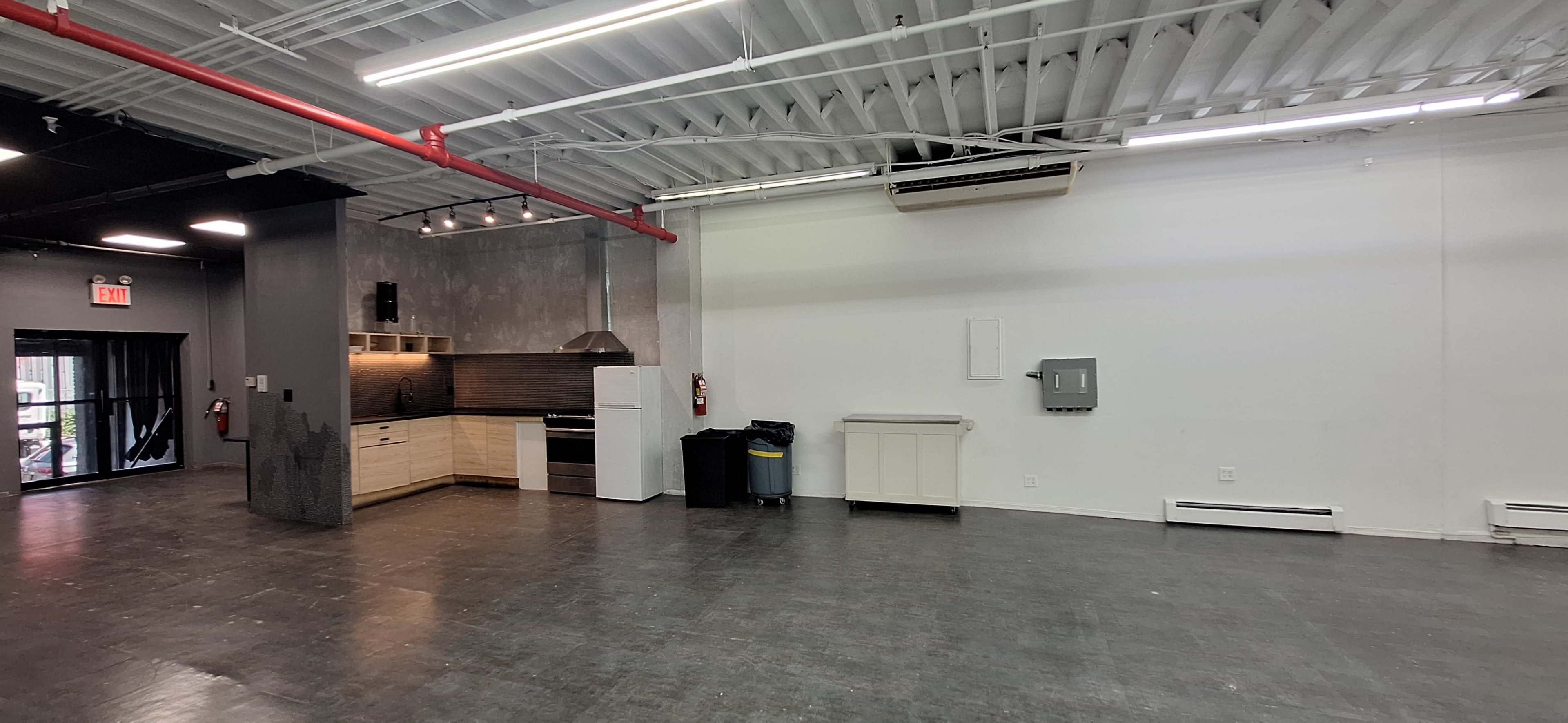 The image shows a spacious, empty room featuring a kitchen area with a stove and cabinets on one side and a blank wall on the opposite side.