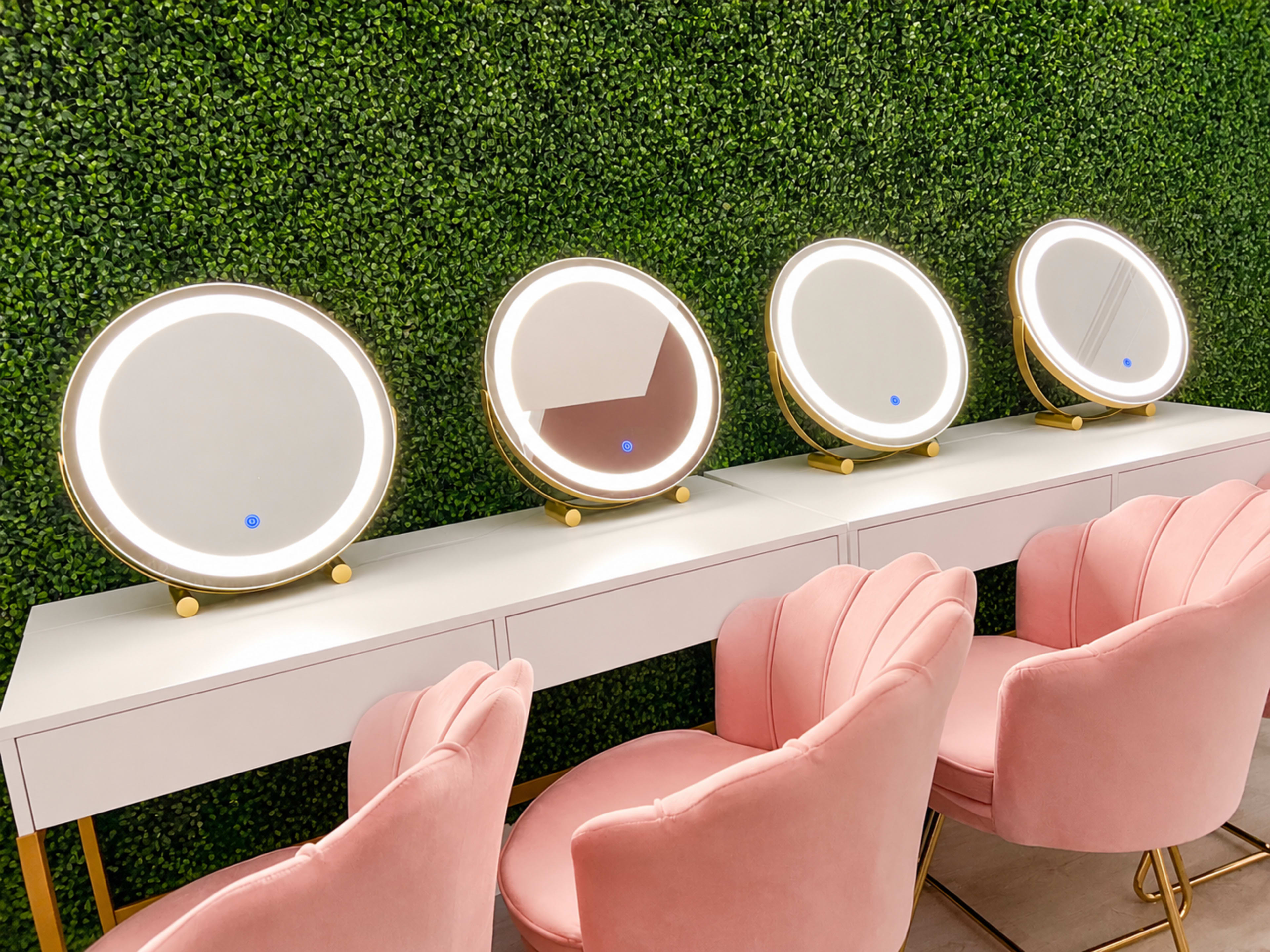 The image shows a row of four illuminated round mirrors set on a white table in front of a green wall, accompanied by pink upholstered chairs.