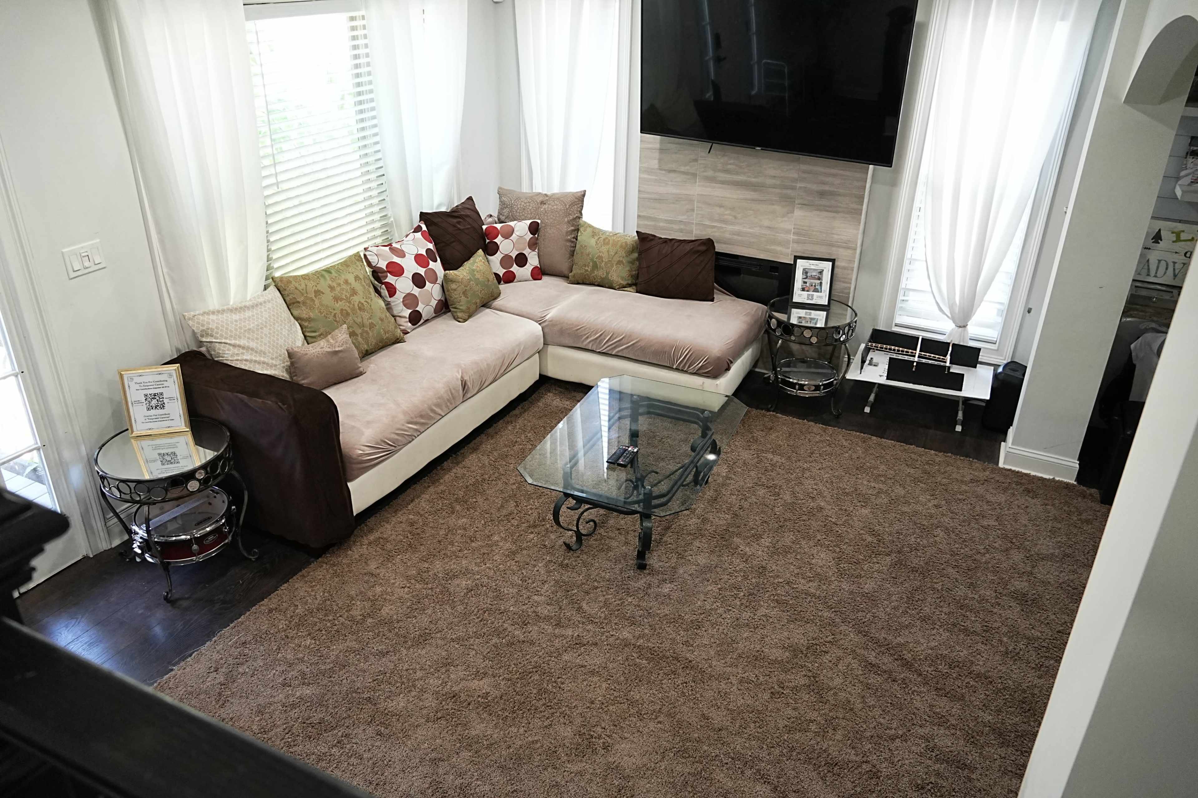 A living room with a sectional sofa, a glass coffee table, and a large area rug, all arranged around a wall-mounted television and large windows with sheer curtains.