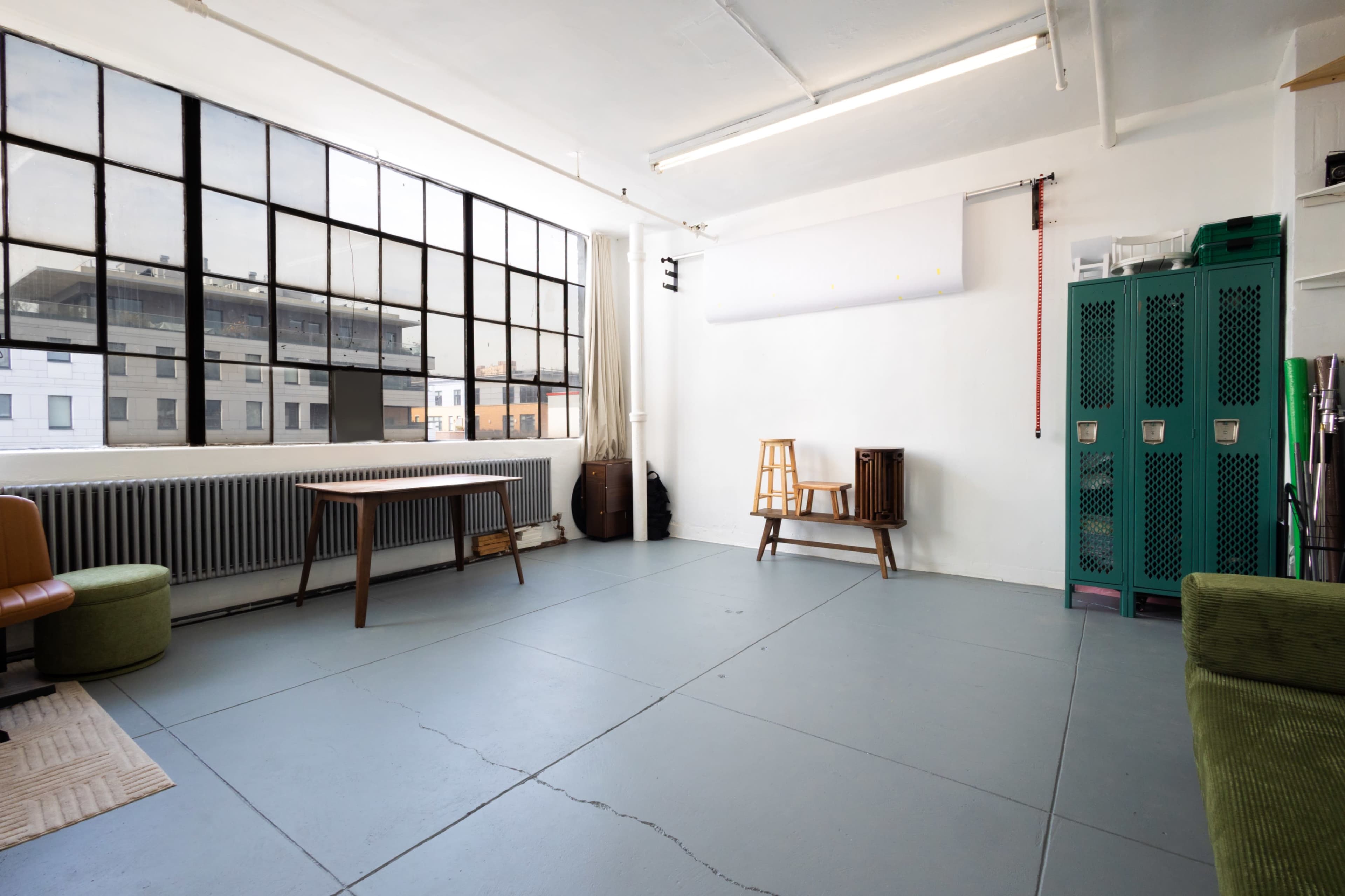 The image shows a spacious, minimalistic room with large windows, a wooden table, a chair, and green lockers against the wall.