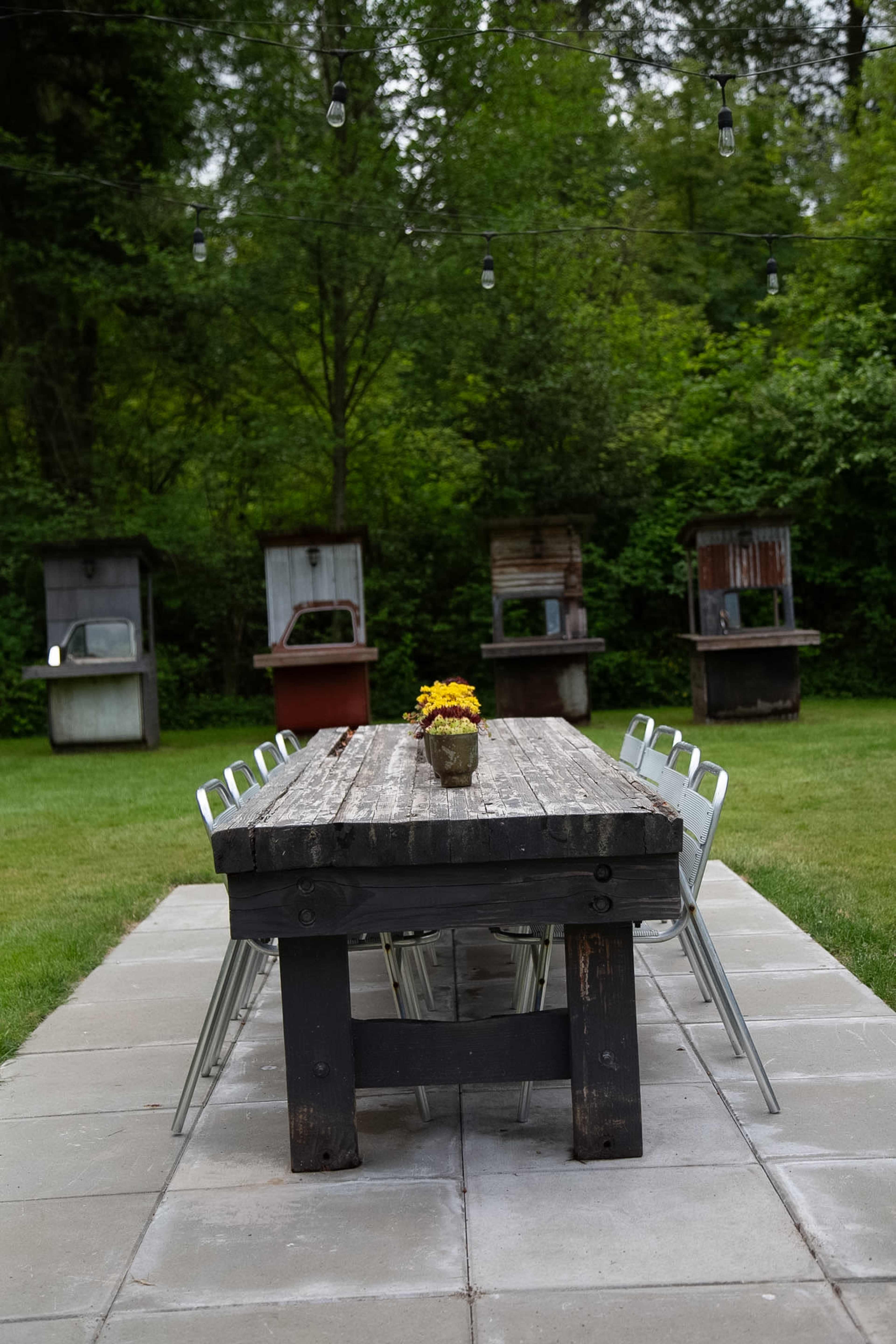 A long, rustic wooden table with metal chairs is set on a stone patio, surrounded by greenery and several old, repurposed displays in the background.