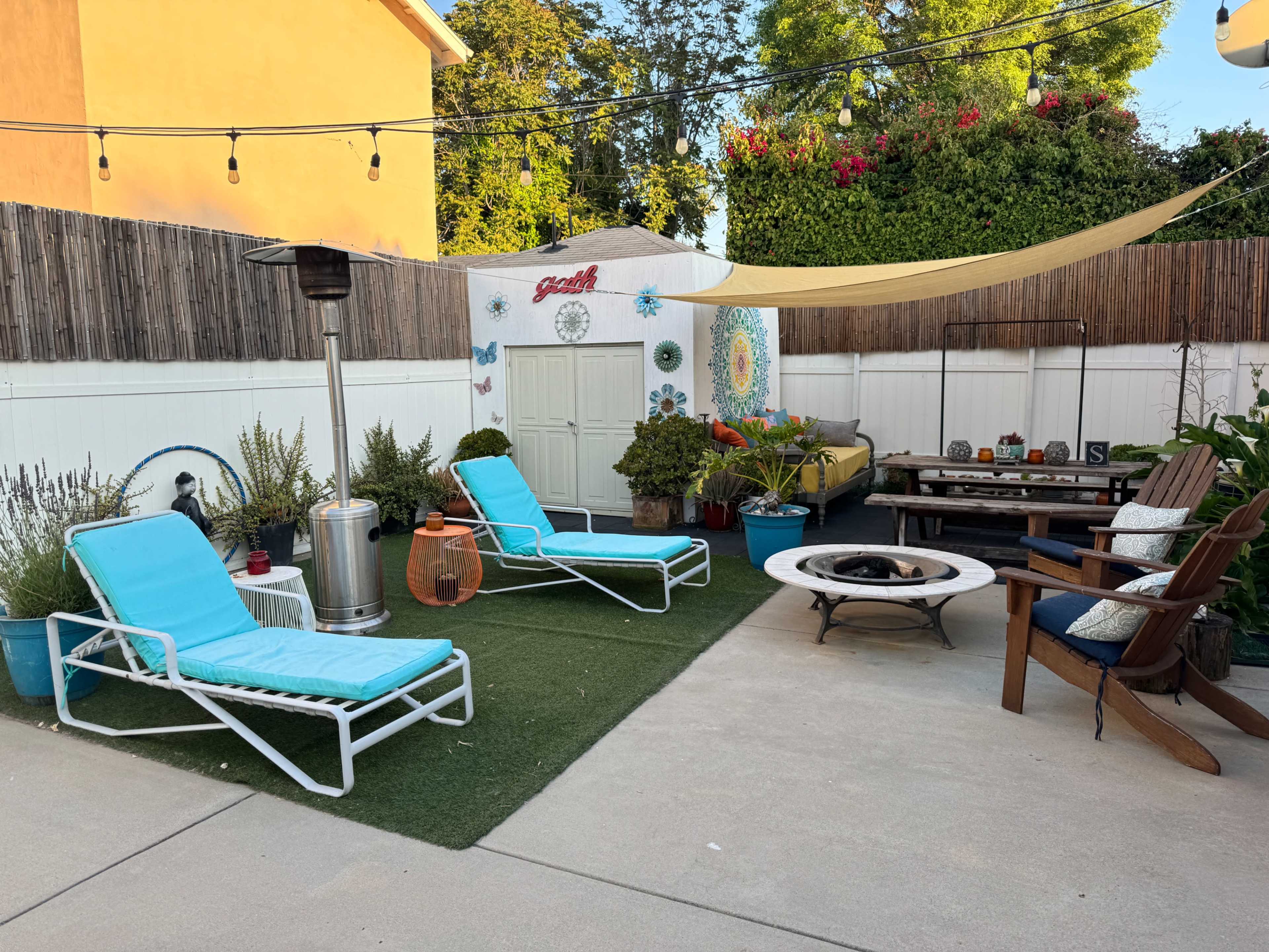 A backyard patio area featuring two light blue lounge chairs, a fire pit, a table with mixed seating, a tall heater, and a decorative shed surrounded by greenery.