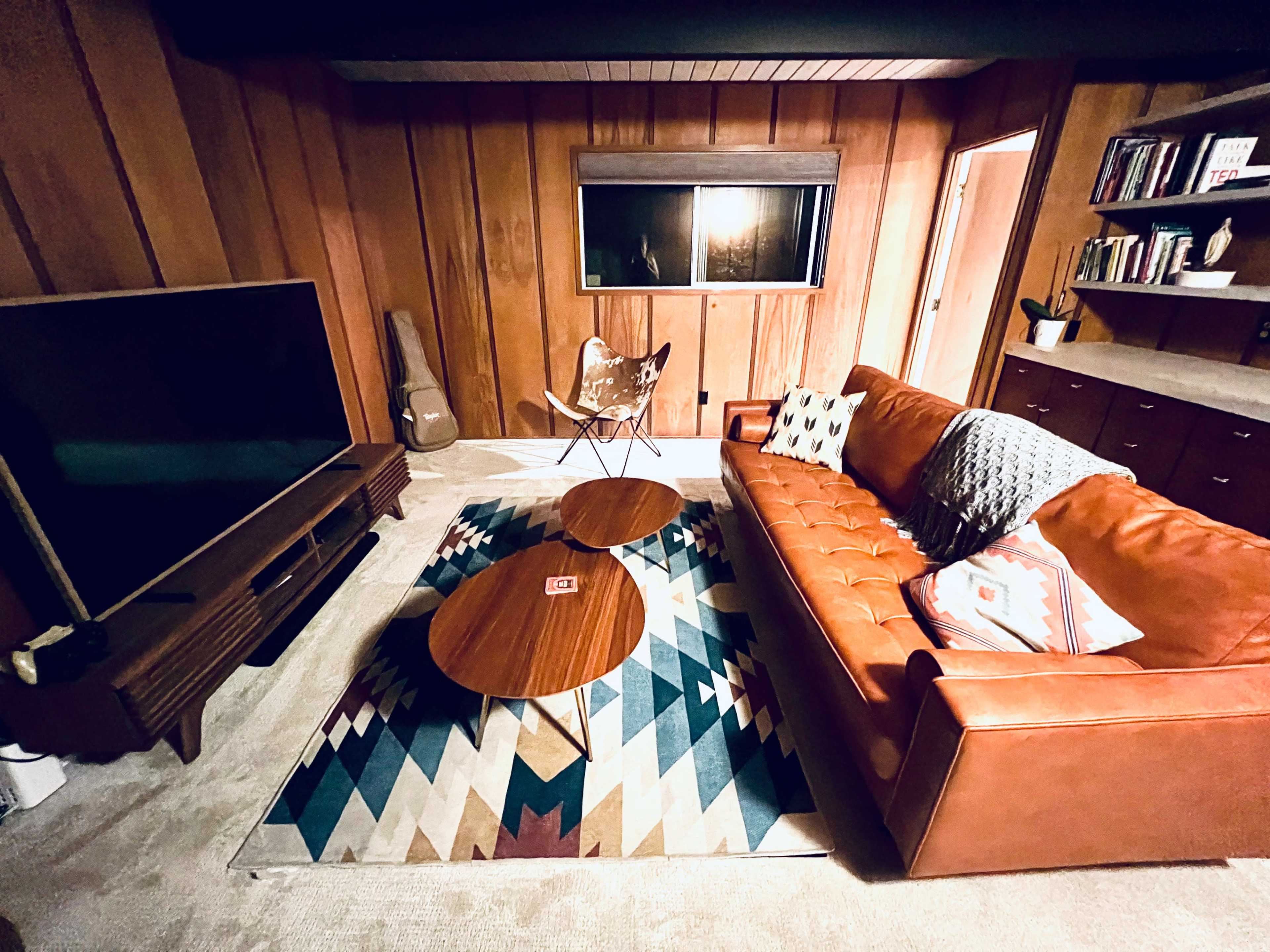 A cozy living room with a brown leather sofa, two wooden coffee tables, a patterned rug, and a large television against wooden-paneled walls.