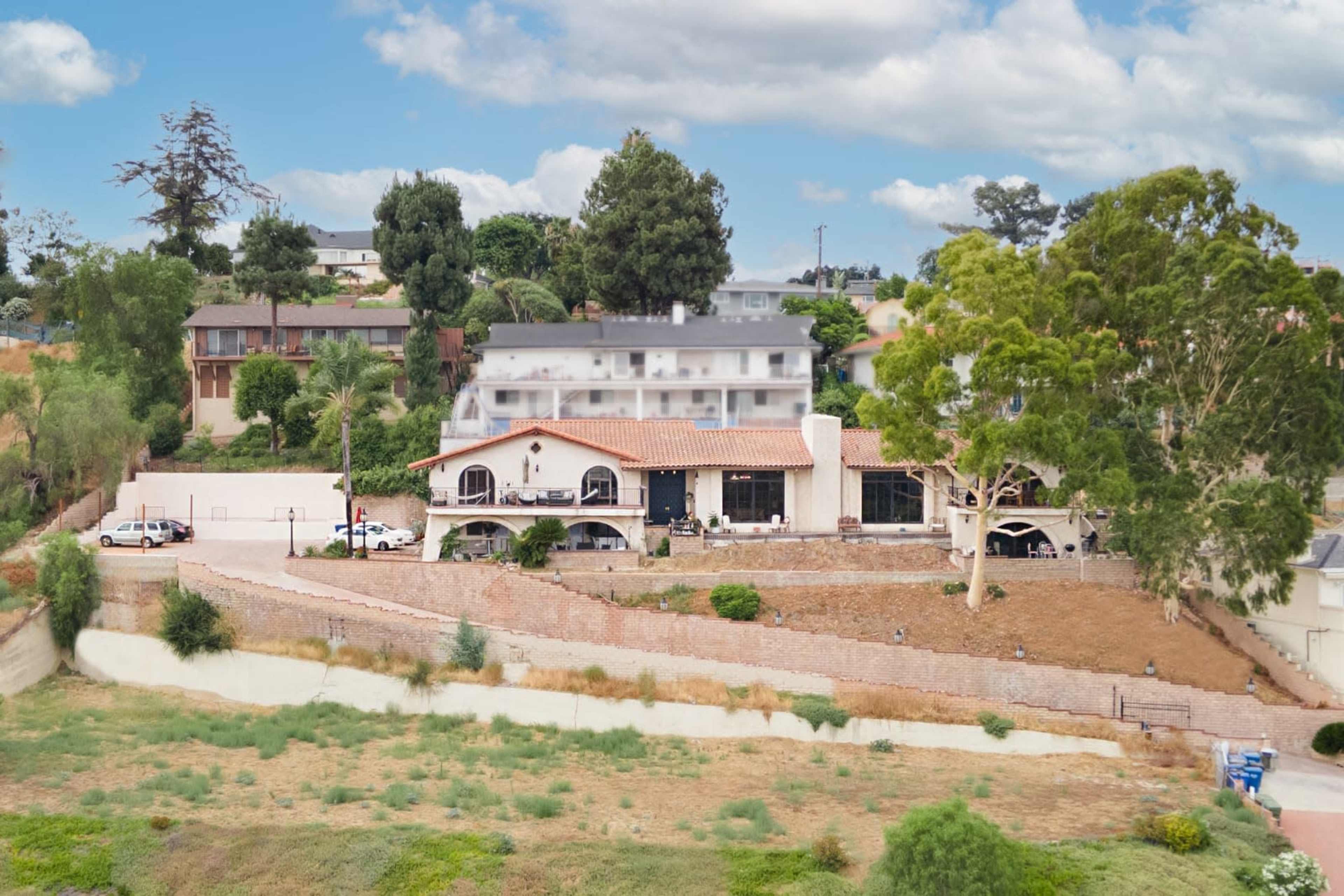 A hillside landscape featuring a house with a tiled roof and several trees, overlooking a grassy area and surrounding residences.