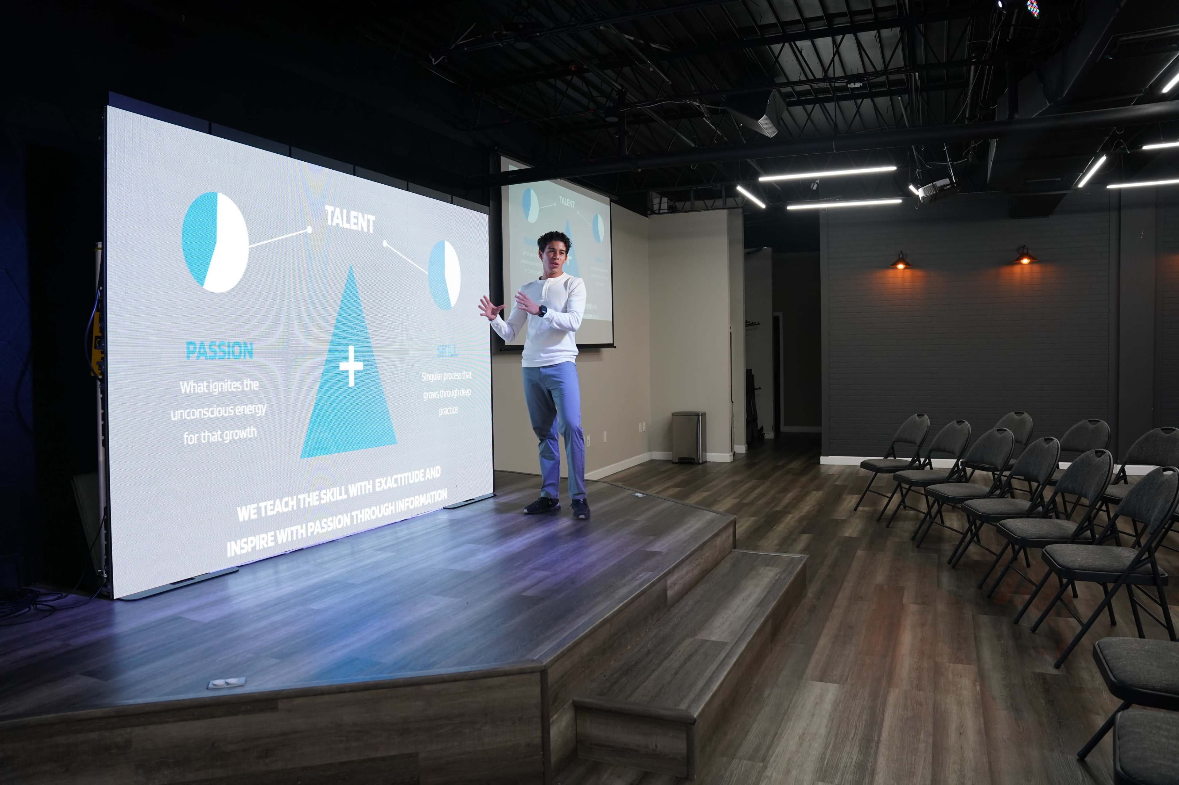 A speaker presents on stage in front of a large screen displaying information about talent and education, while chairs are arranged for an audience.