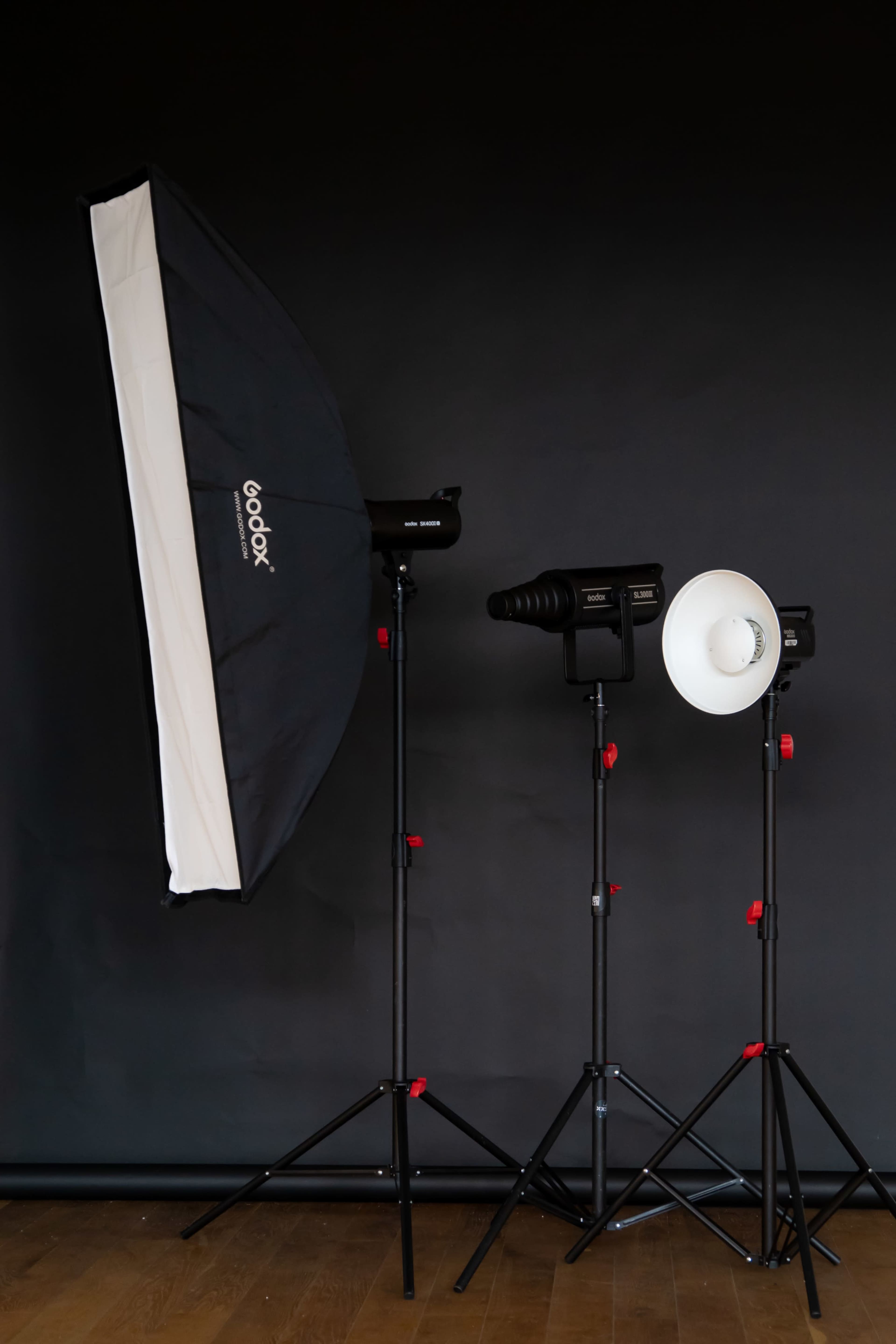 The image shows three photography lighting equipment setups, including a large softbox and two studio lights, on adjustable tripods against a black background.