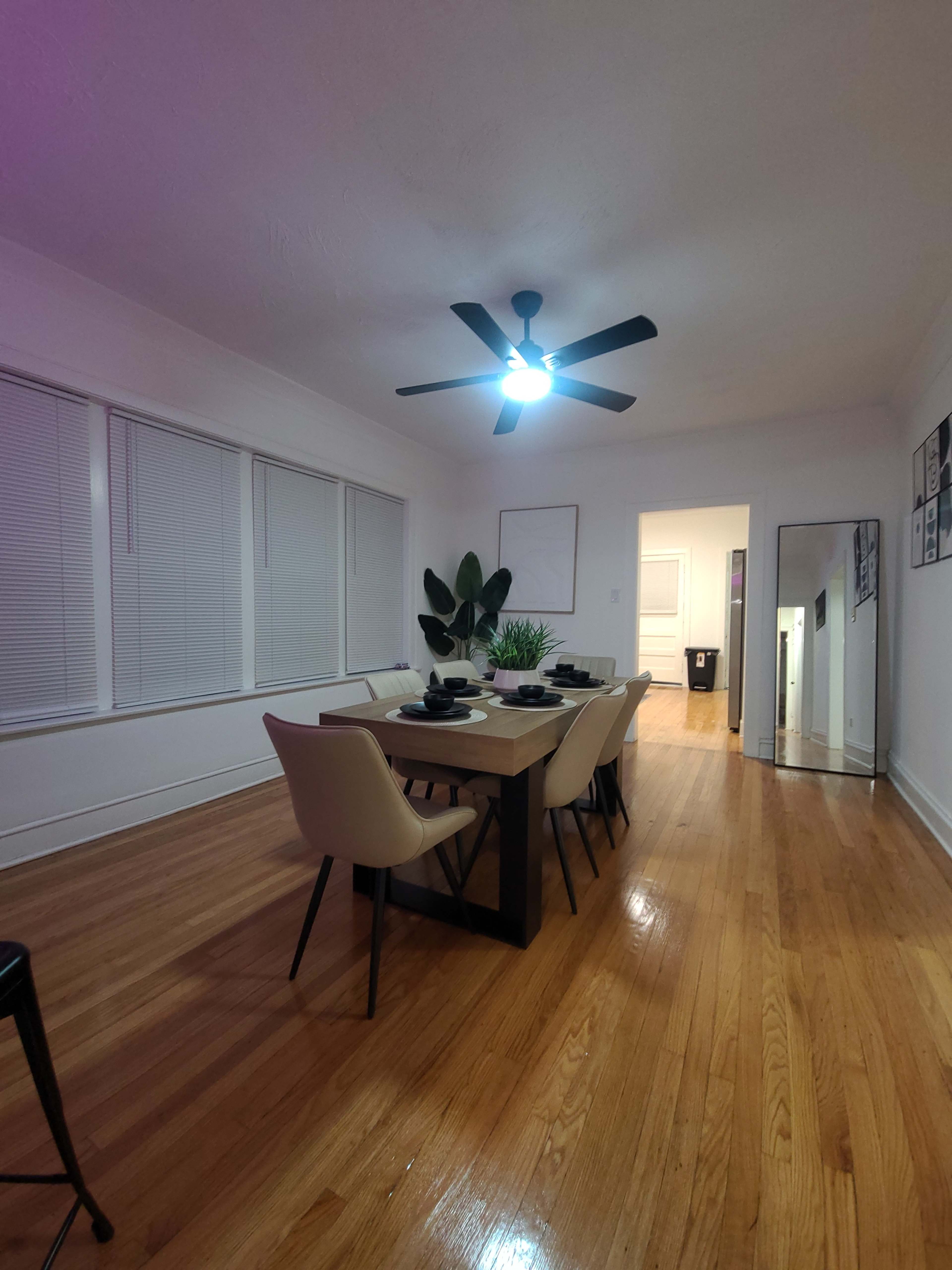 A dining room features a long wooden table set for a meal, surrounded by chairs, with a ceiling fan above and a potted plant in the corner.