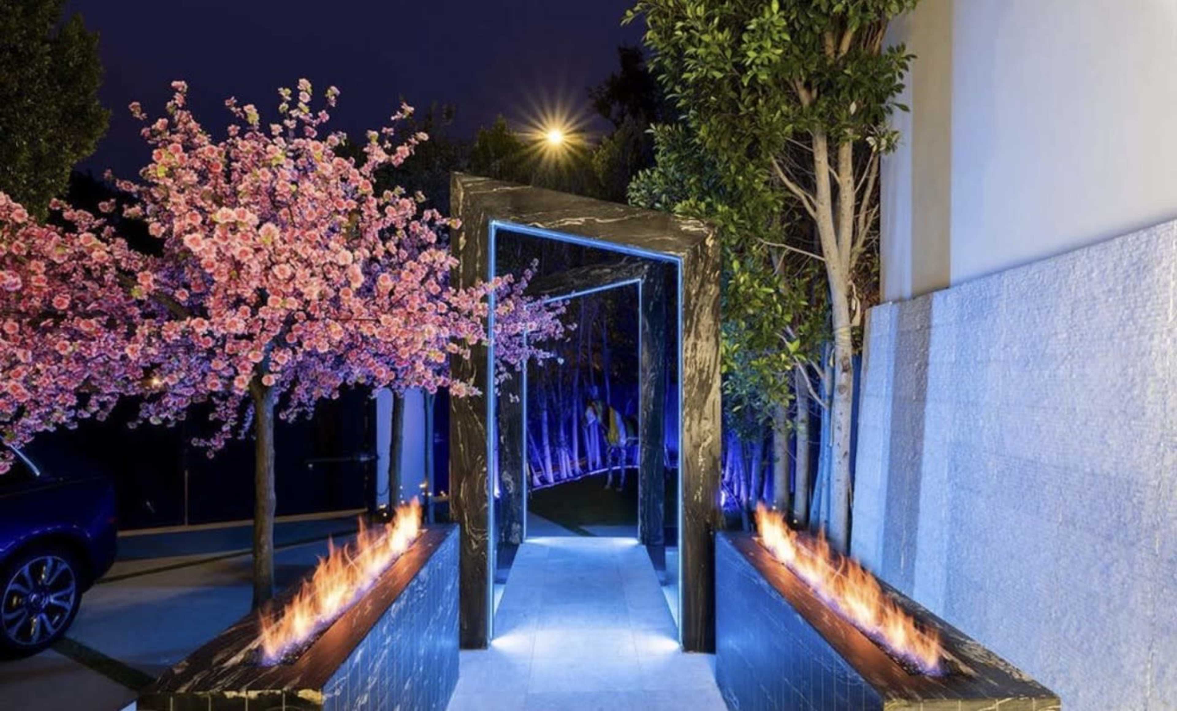 The scene features a stylish entranceway with a black arch surrounded by illuminated trees and flaming water features.