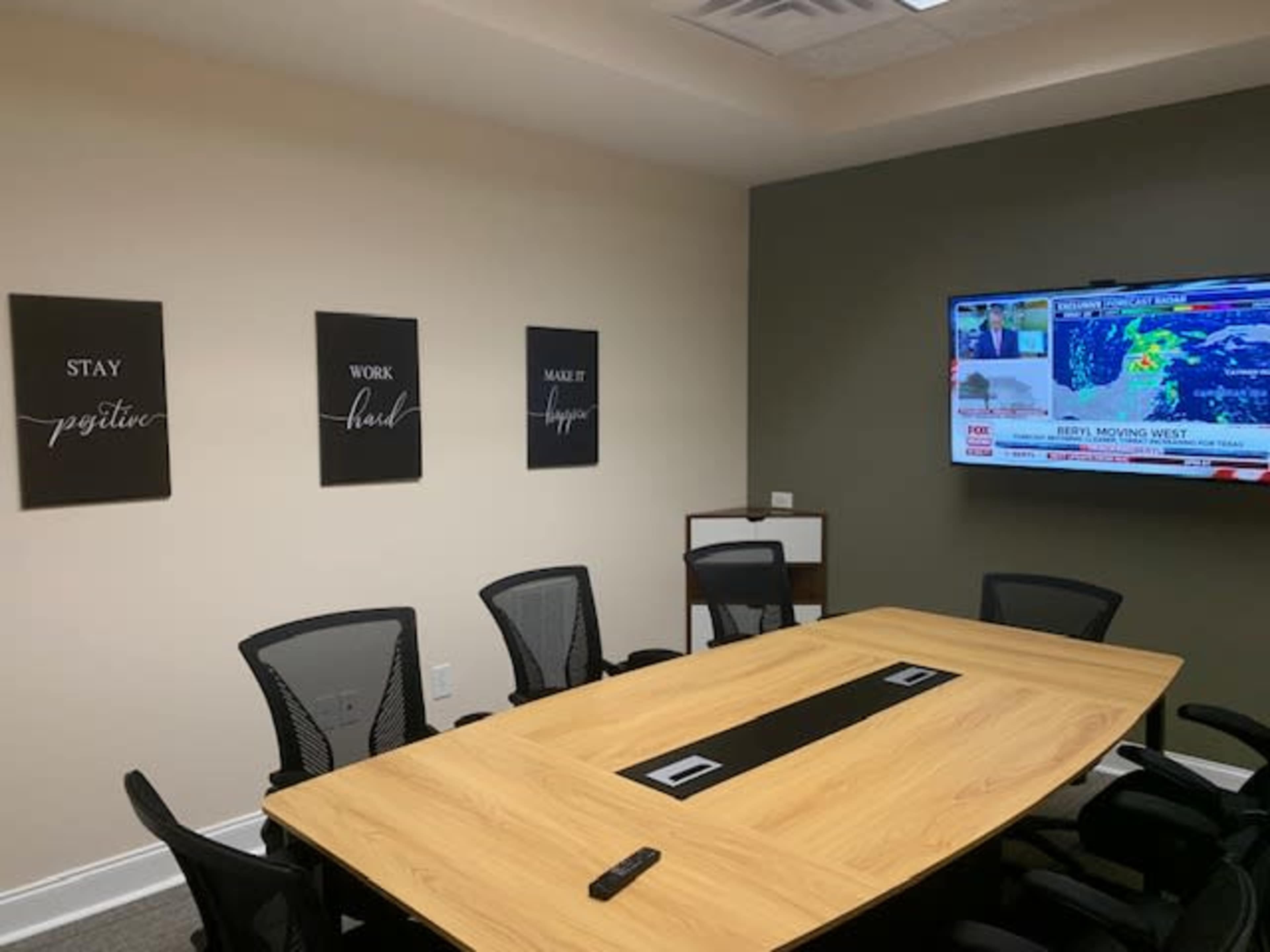 A conference room features a large wooden table surrounded by black chairs and motivational posters on the wall, with a television displaying a news broadcast.