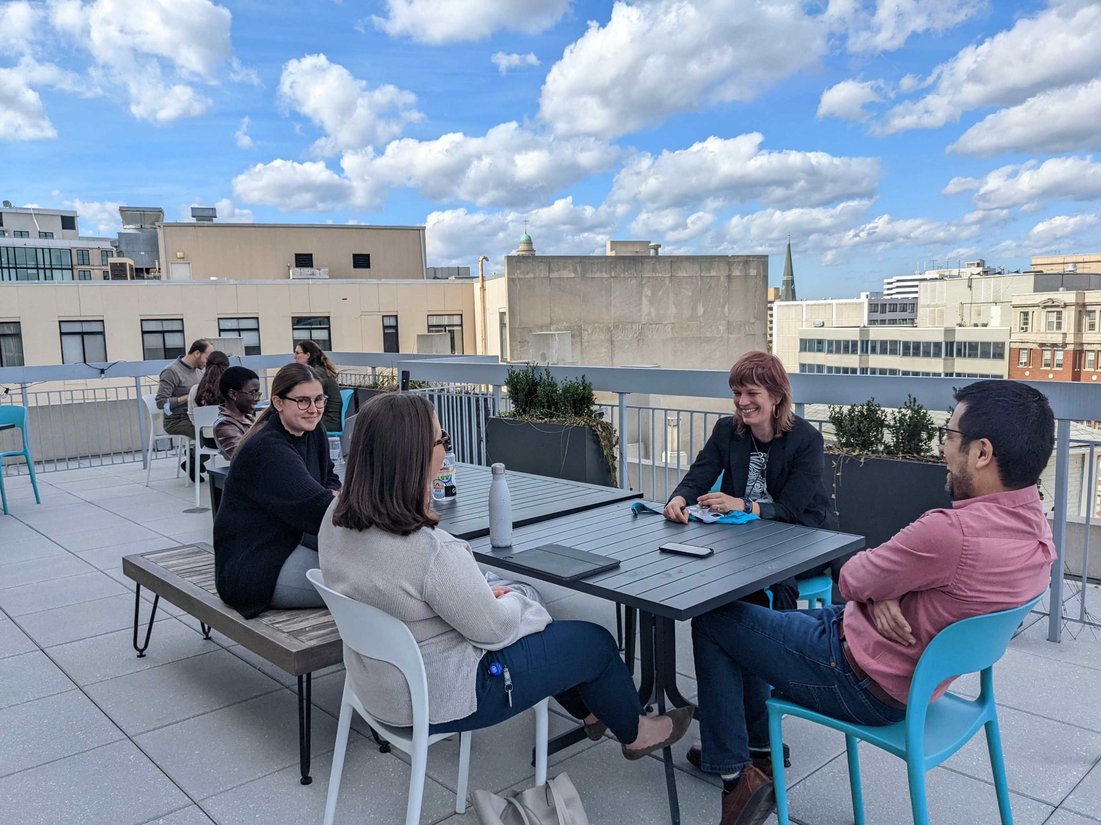 A group of five people sit around a table on a rooftop, discussing while surrounded by urban buildings and a partly cloudy sky.