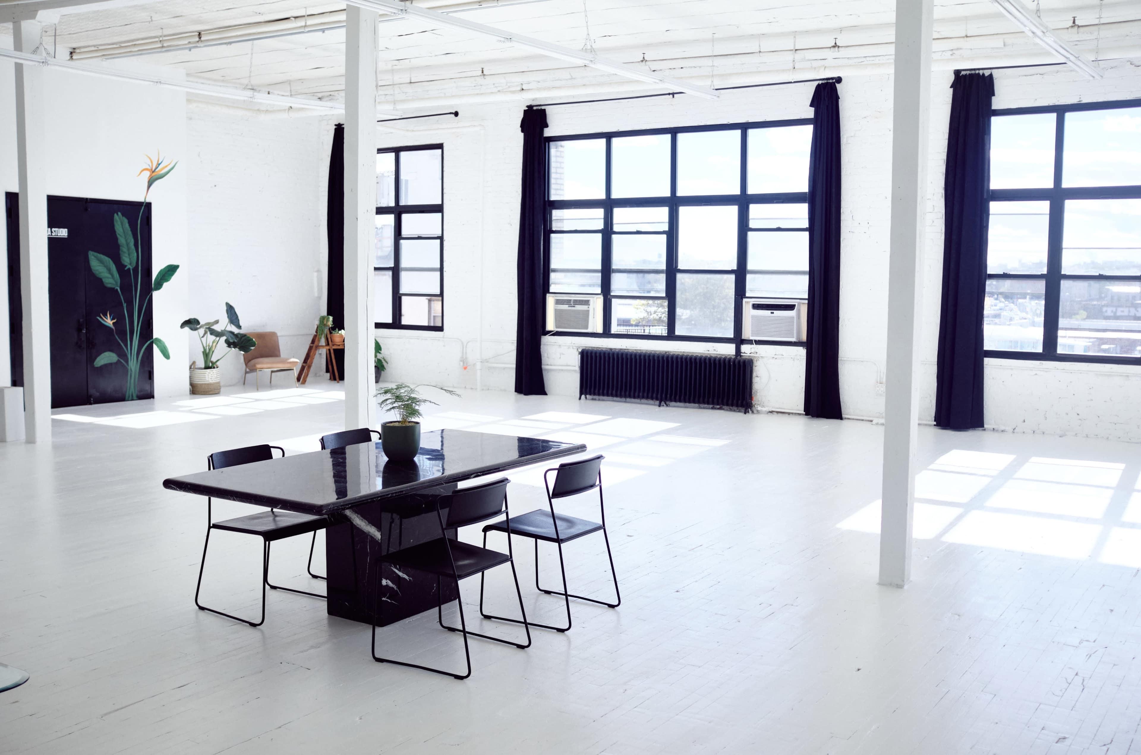 A spacious, minimalistic room with large windows, a black table surrounded by chairs, and a plant near a corner.