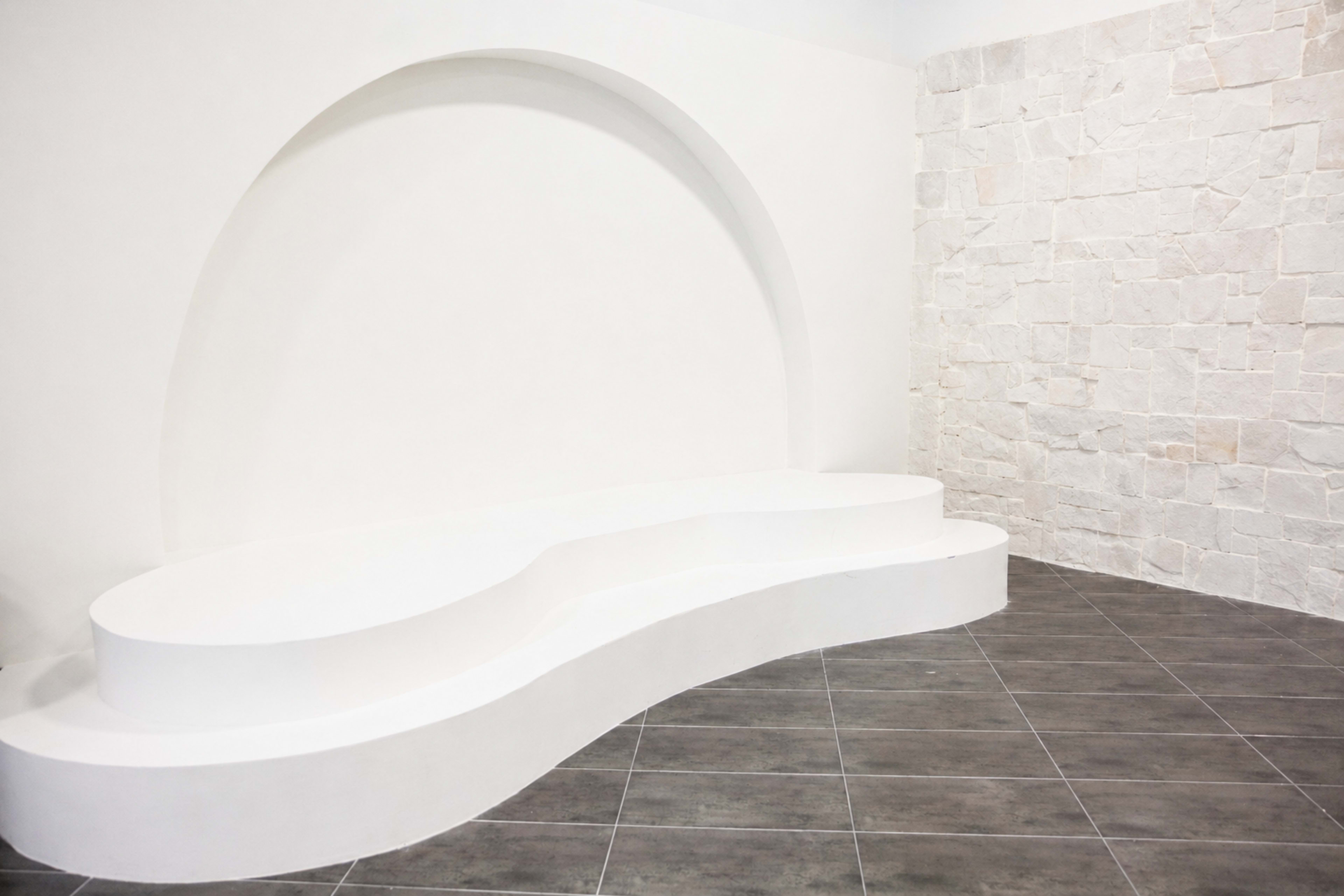 The image shows a minimalist interior featuring a curved white bench against a light stone wall with gray tile flooring.