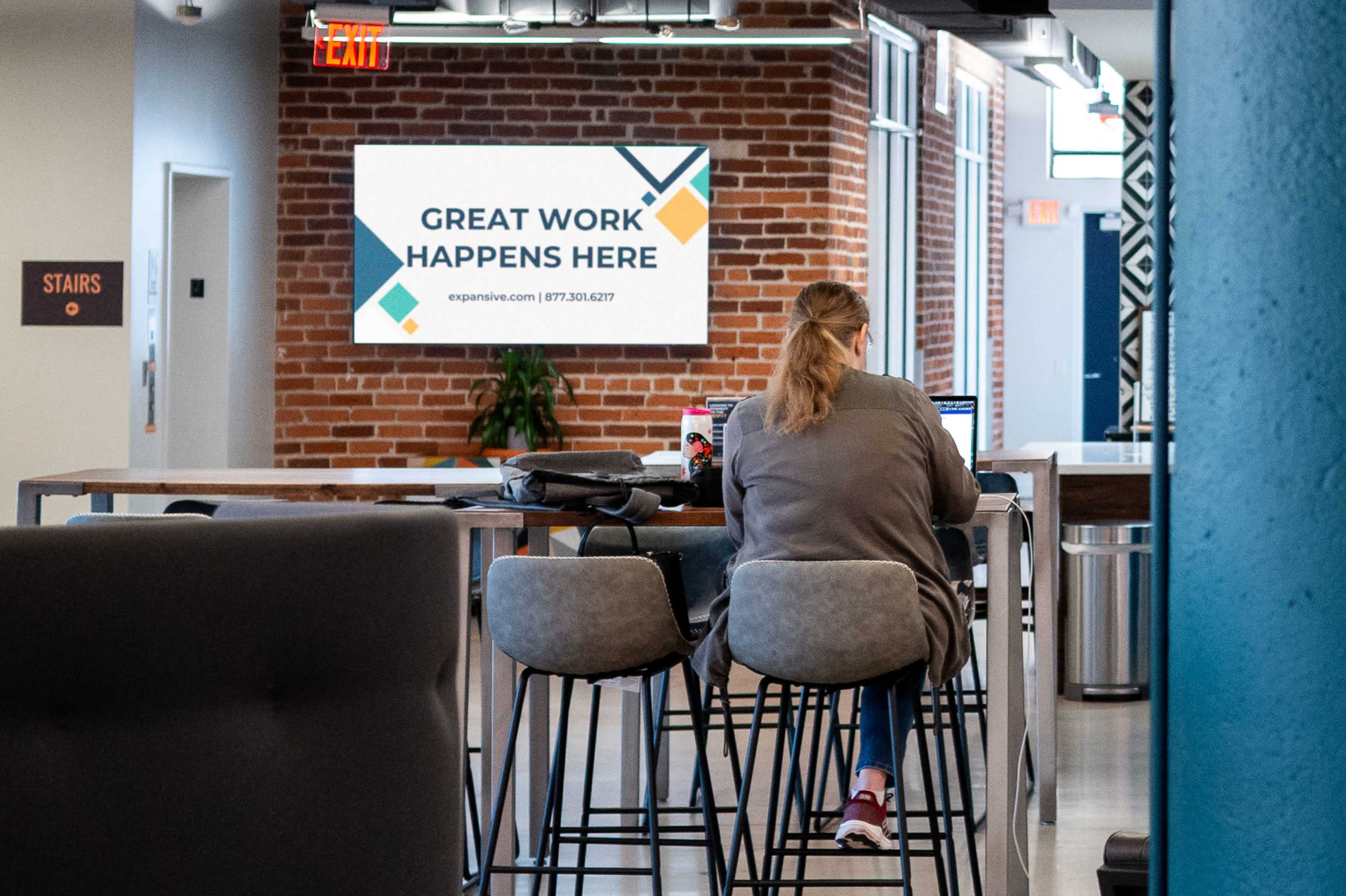 A person sits at a high table working on a laptop in a modern office setting, with a monitor displaying "GREAT WORK HAPPENS HERE" in the background.