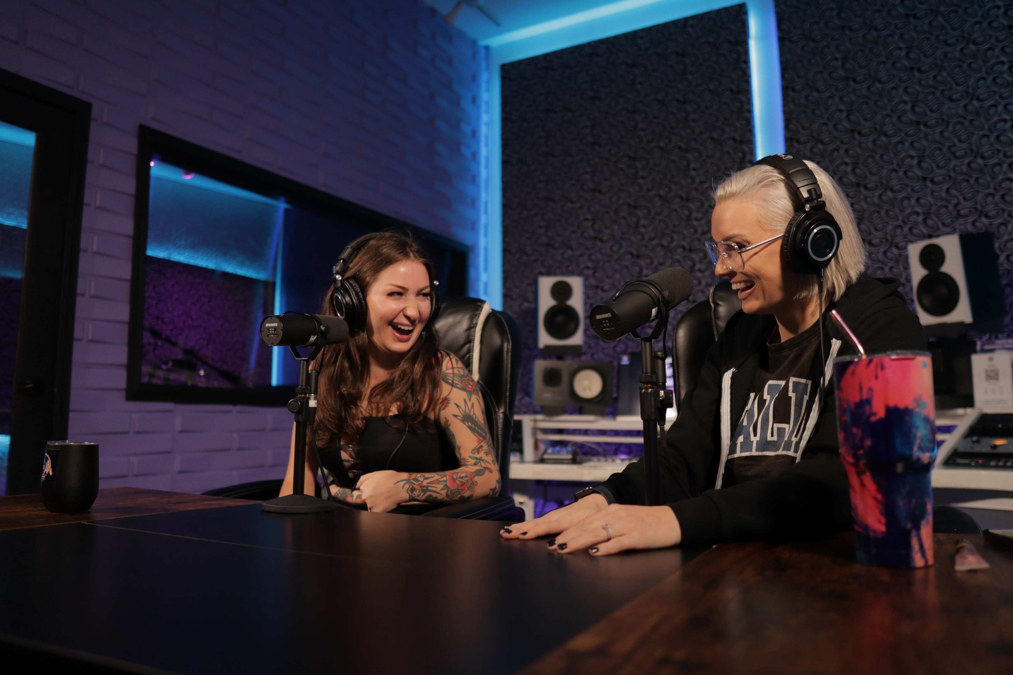 Two women are seated at a table in a recording studio, both wearing headphones and laughing while speaking into microphones.