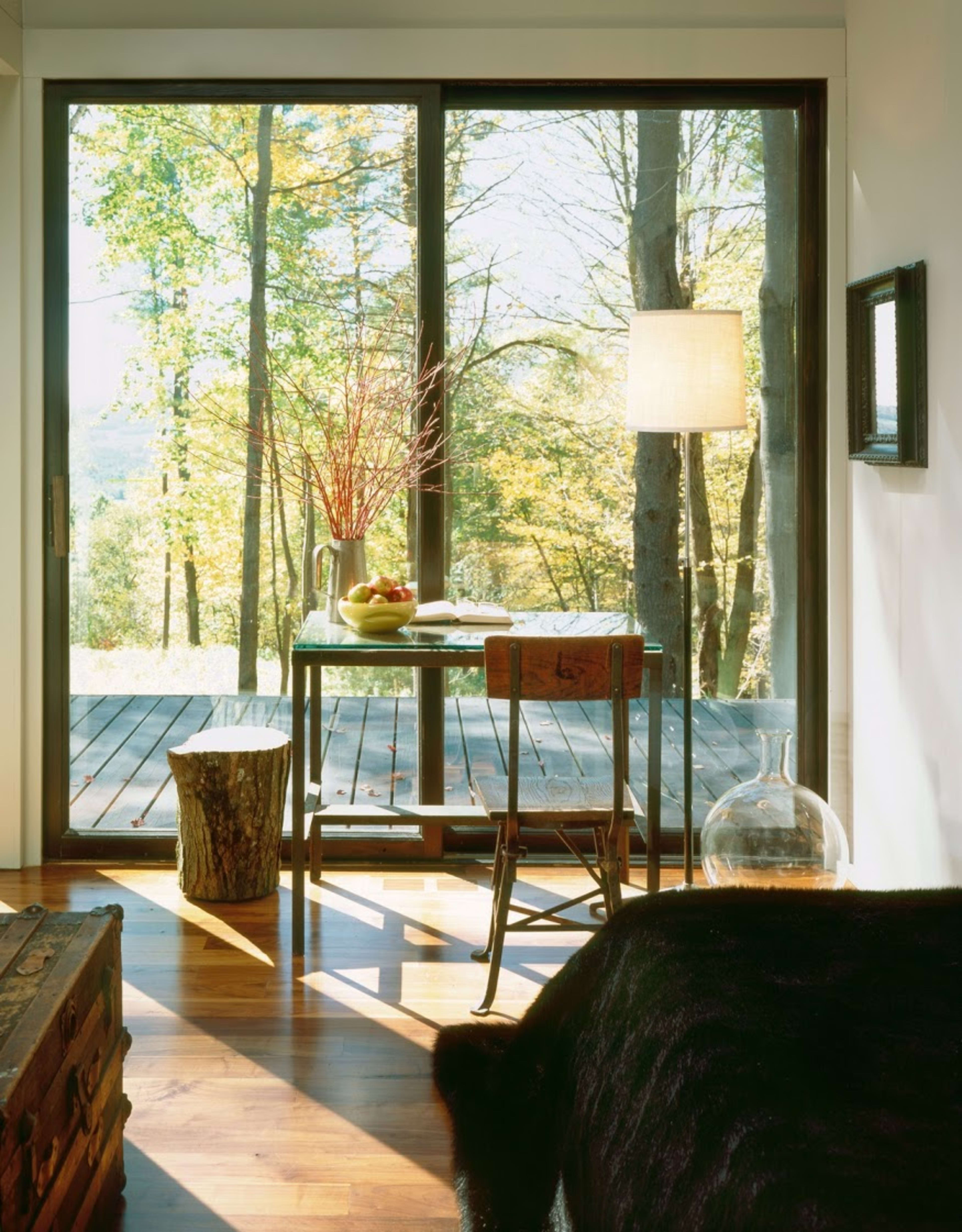 A simple wooden table with a bowl of fruit is positioned near a large window that overlooks a forested area.