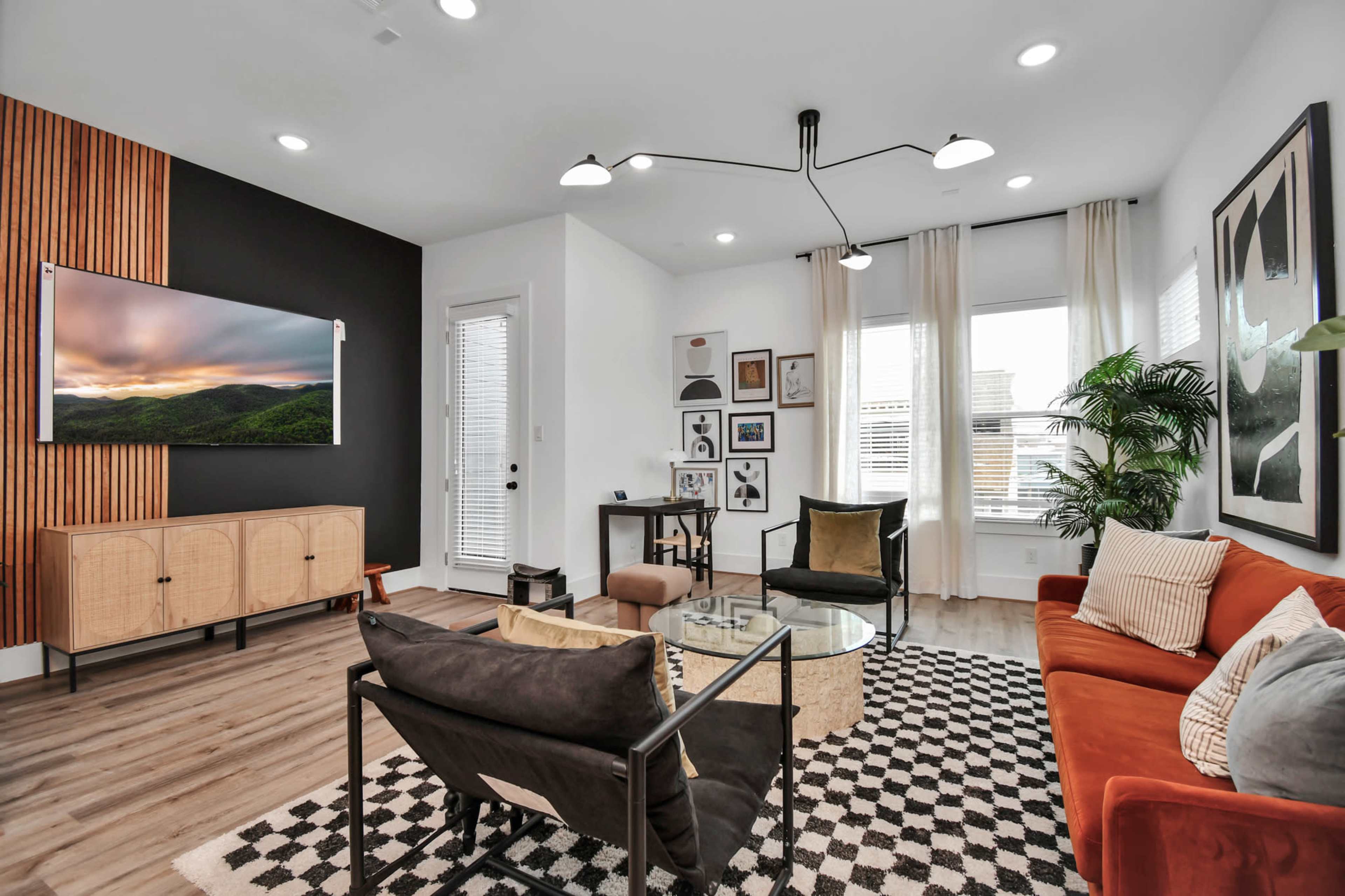 The image shows a modern living room with a large TV mounted on the wall, a glass coffee table, an orange sofa, and a patterned rug.