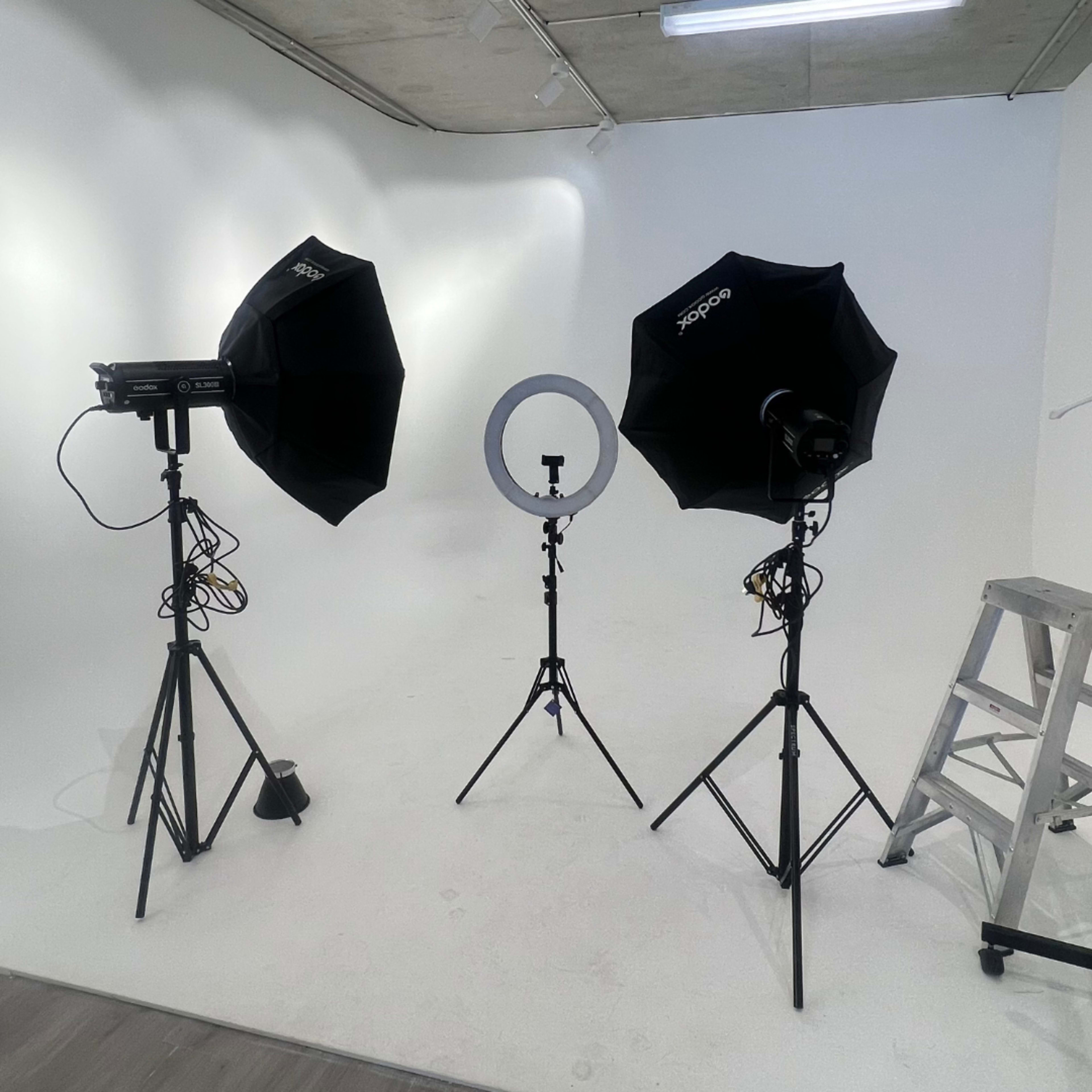The image shows a photography studio setup with two softboxes, a ring light, and a ladder positioned against a plain white backdrop.