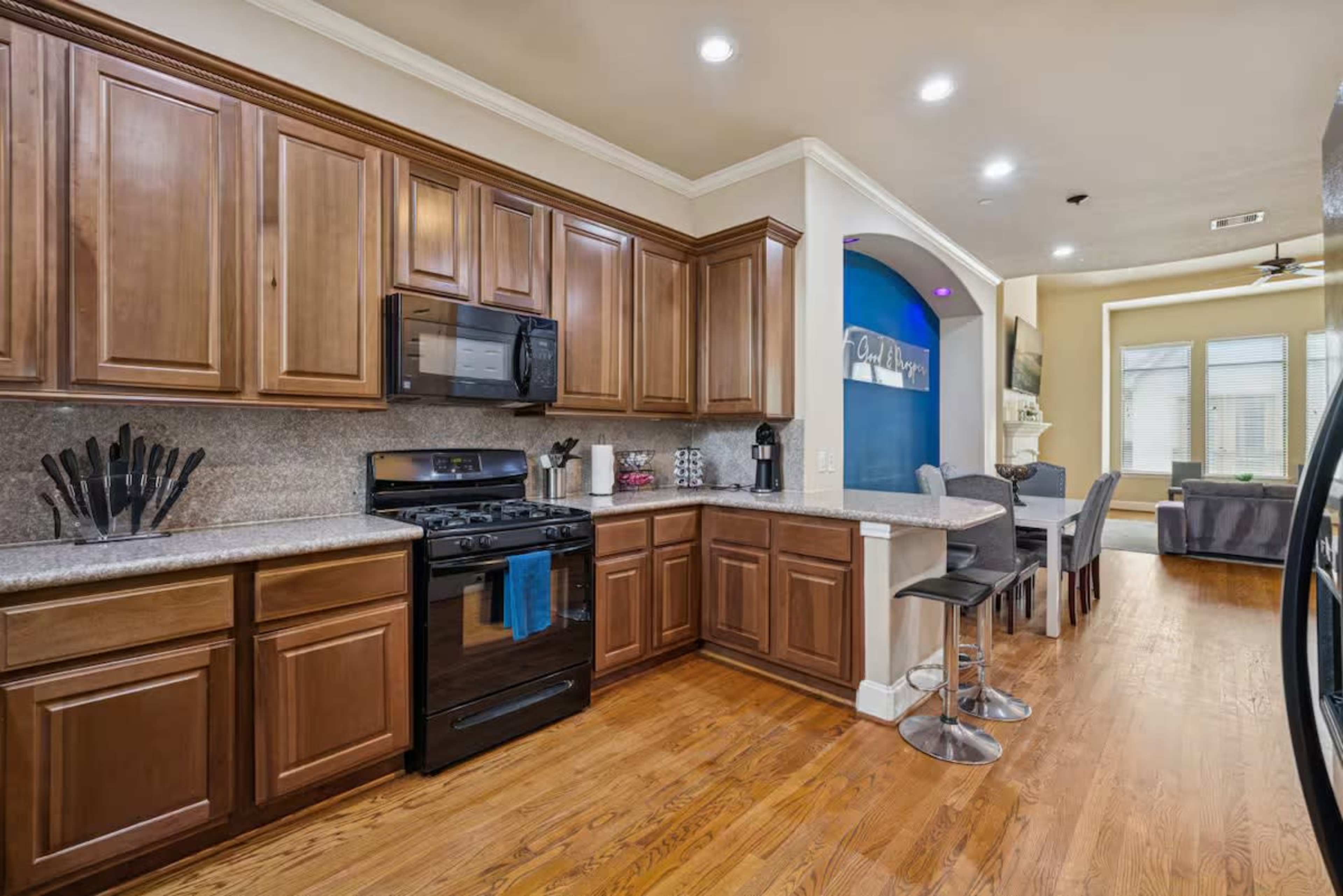 The image shows a modern kitchen with wooden cabinetry, a gas stove, and a breakfast bar with stools, adjacent to a living area.