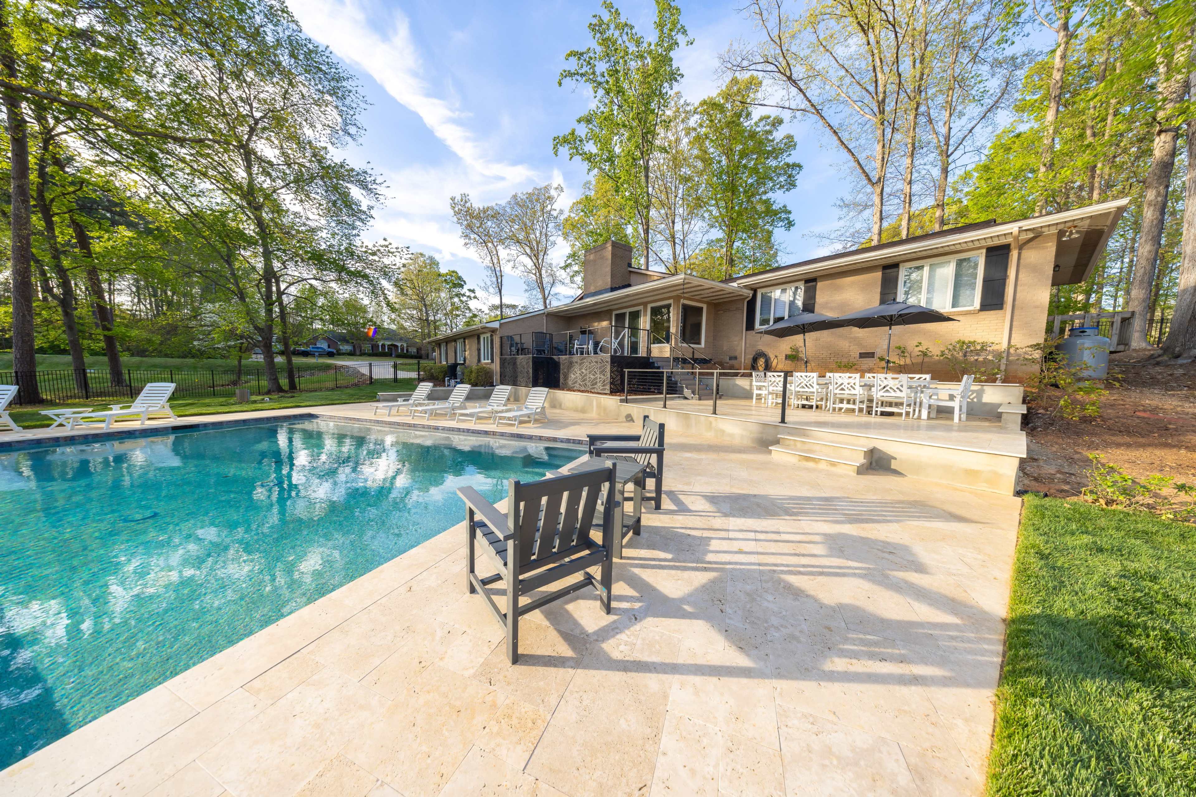 A backyard features a rectangular swimming pool with lounge chairs and a house in the background surrounded by trees.