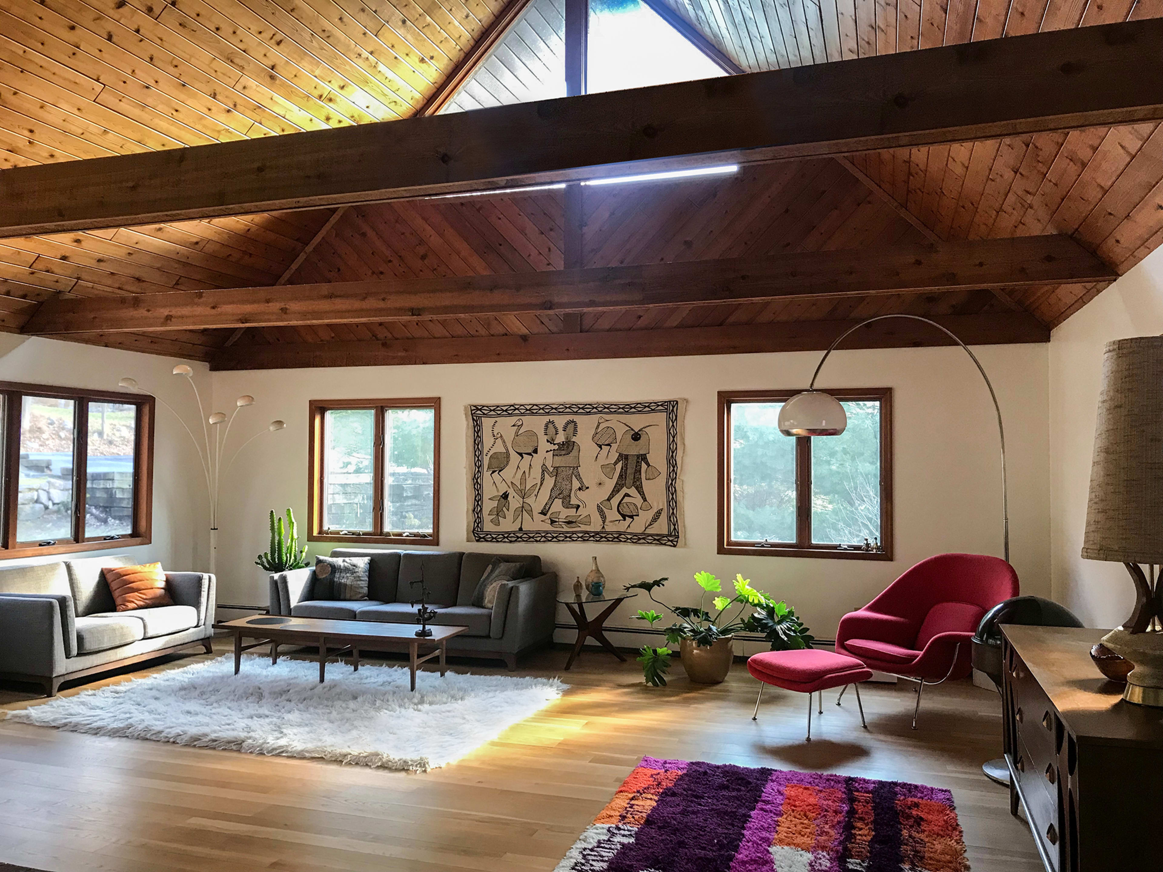 A spacious living room features a high wooden ceiling, large windows, a modern sofa set, and decorative plants.