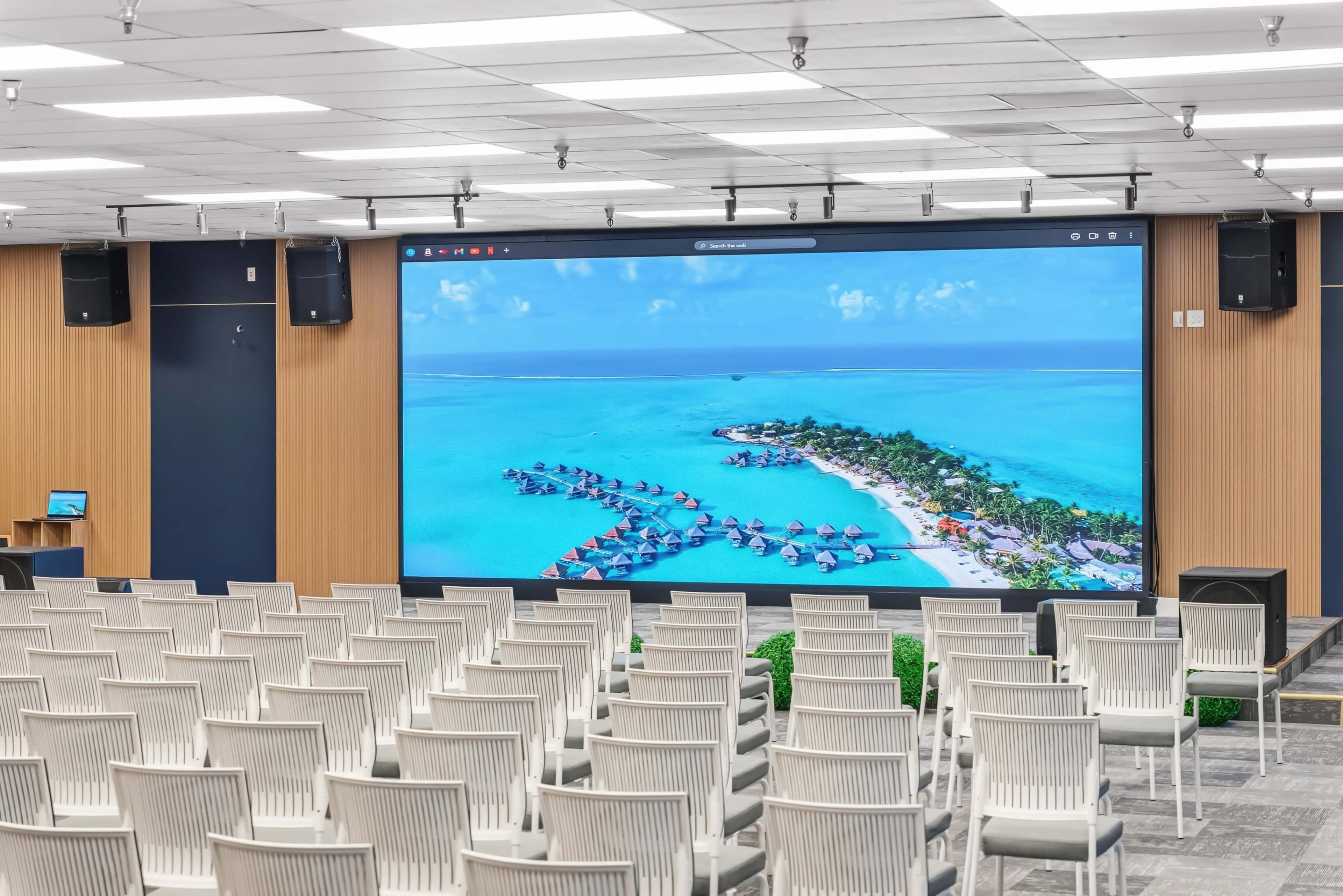 A large conference room features a wall-sized screen displaying an aerial view of a tropical resort with overwater bungalows surrounded by turquoise water, while rows of empty white chairs face the screen.