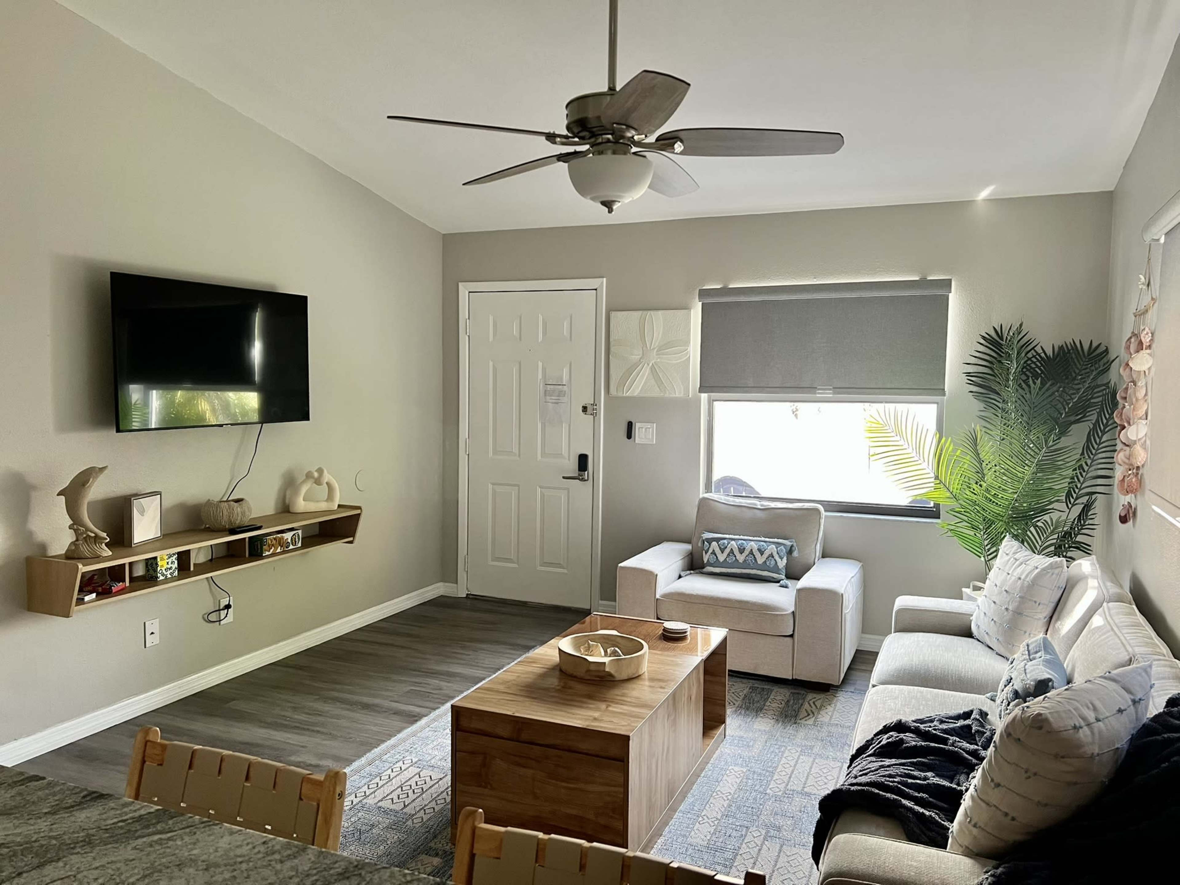 The image shows a modern living room with a flat-screen TV mounted on the wall, a coffee table in the center, a couch, and a large window featuring a shaded view.