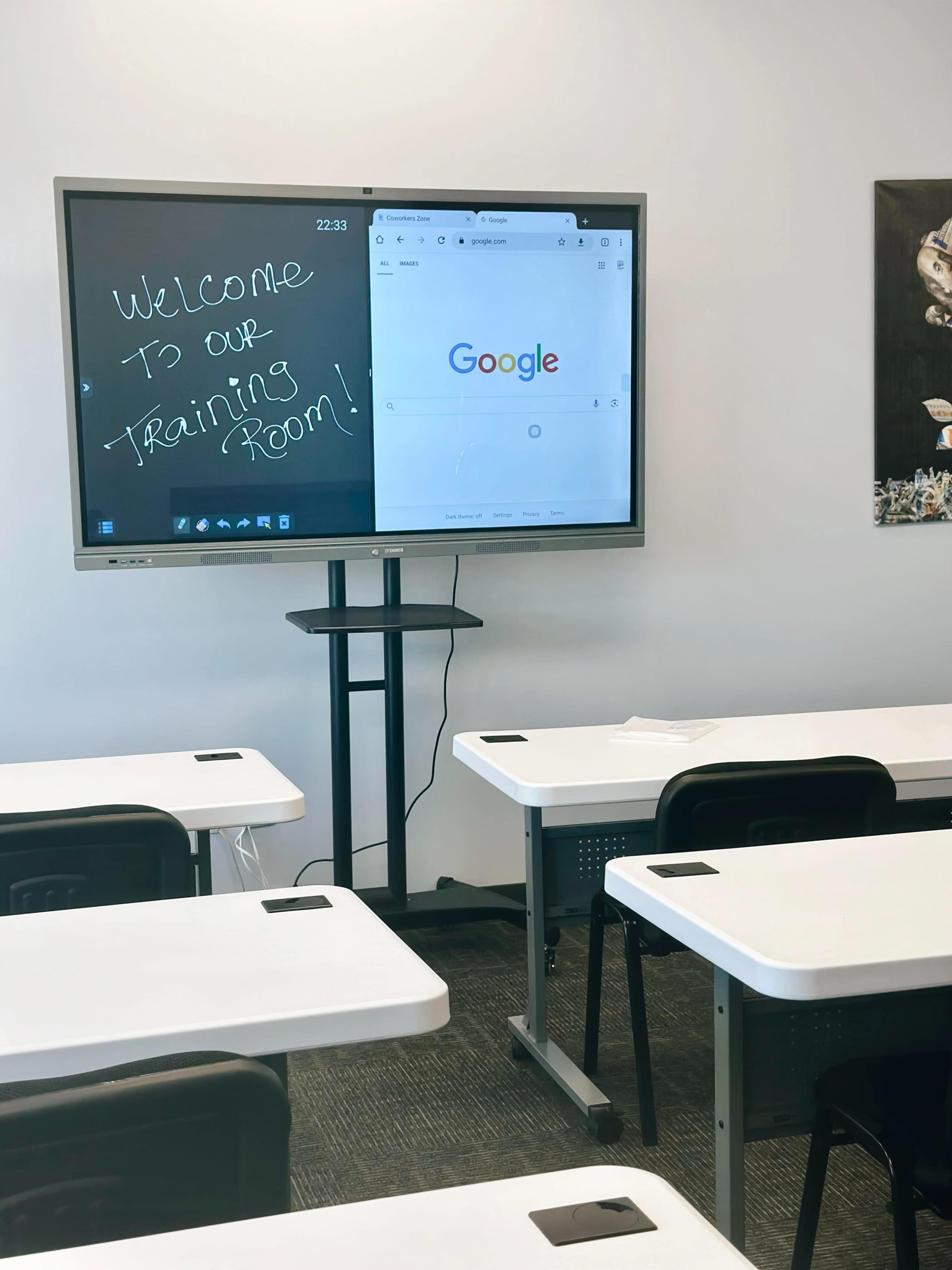 A digital display in a classroom shows a welcome message alongside a Google search page, with empty desks arranged in front.