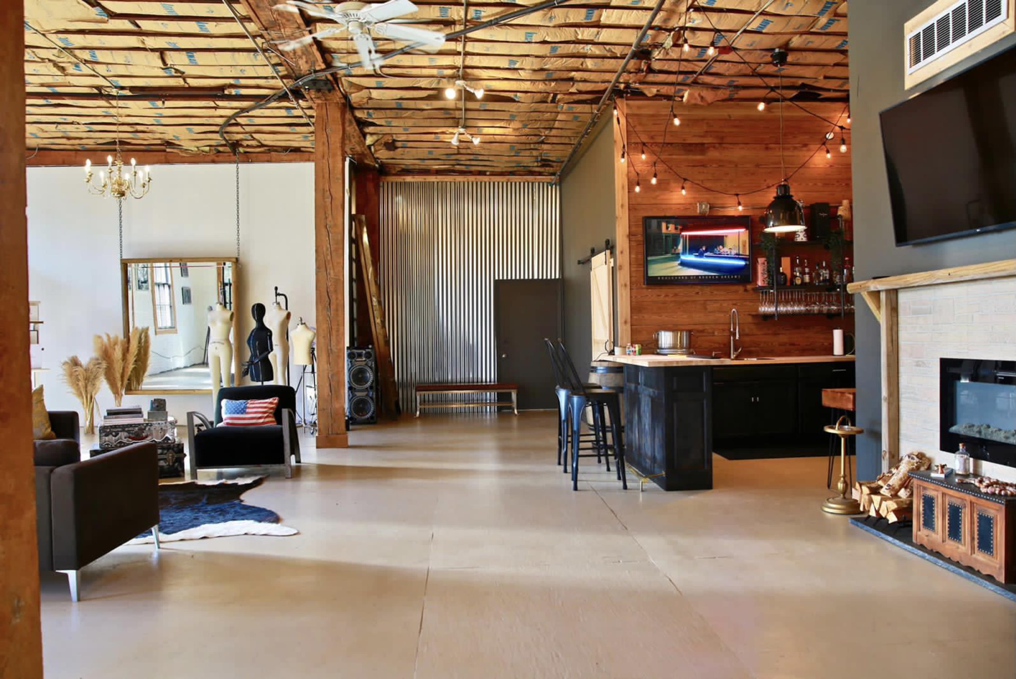 A spacious industrial-style interior featuring exposed beams, a kitchen area, a seating arrangement with dark furniture, and a decor that includes a full-length mirror and artwork.