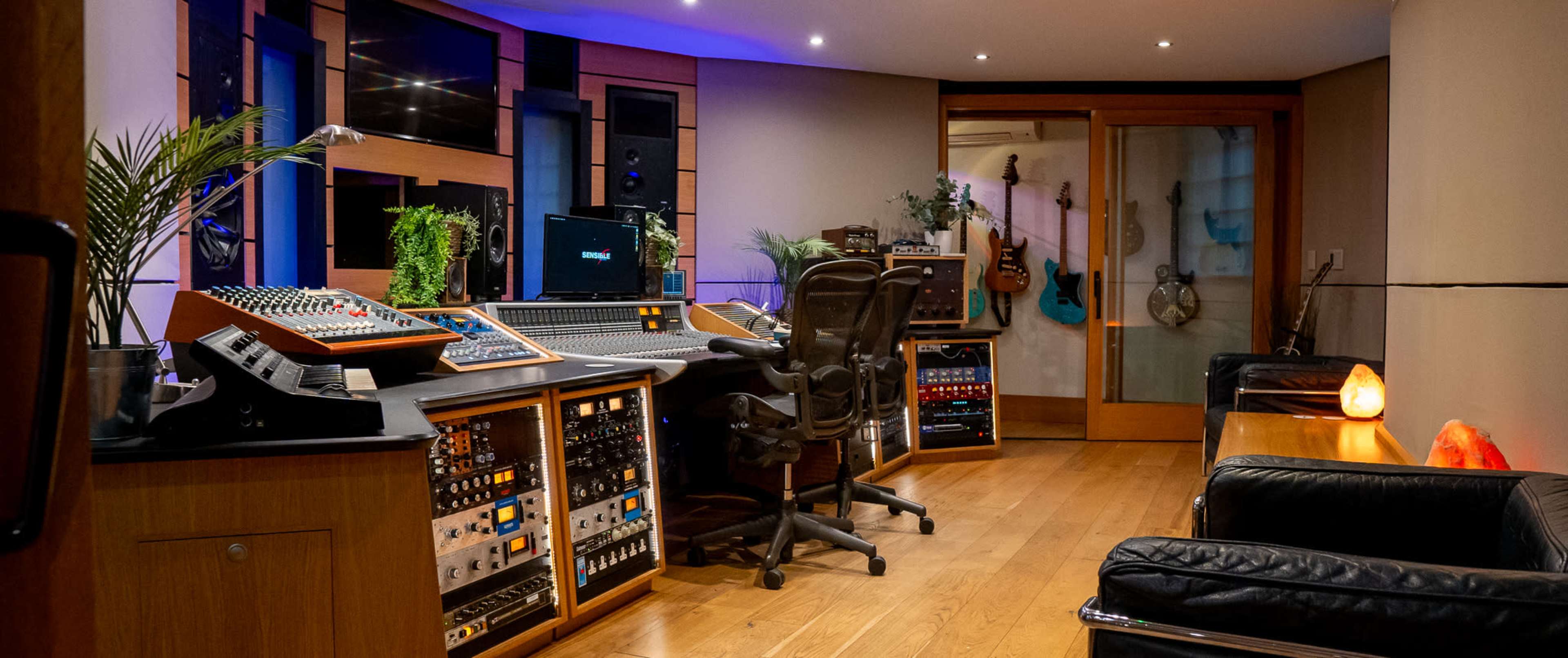 The image shows a music studio featuring a large mixing console, various audio equipment, and musical instruments arranged in a modern interior with warm wood flooring.