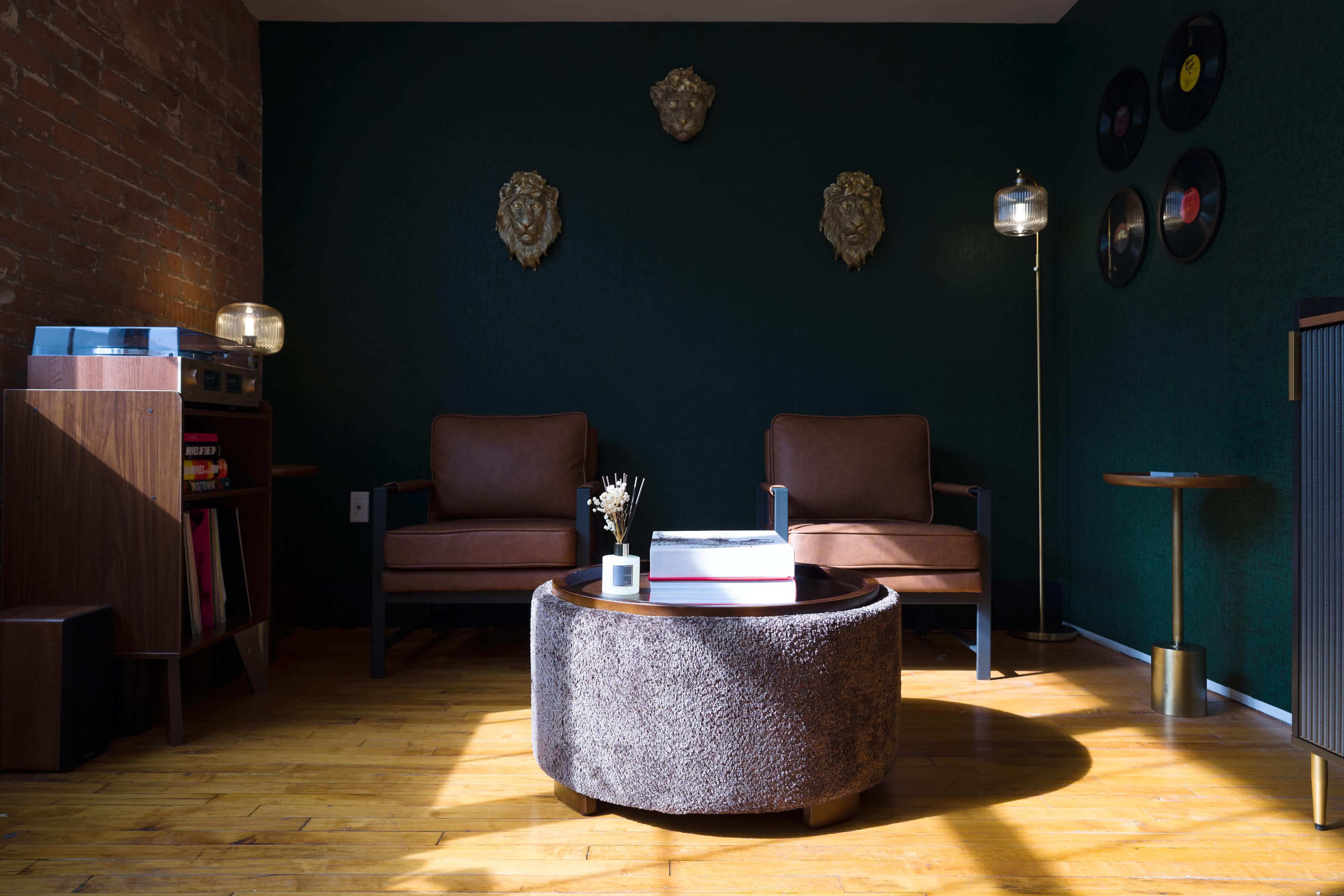 The image shows a cozy living room featuring two brown leather chairs, a round coffee table, and decorative wall accents on a dark green background.