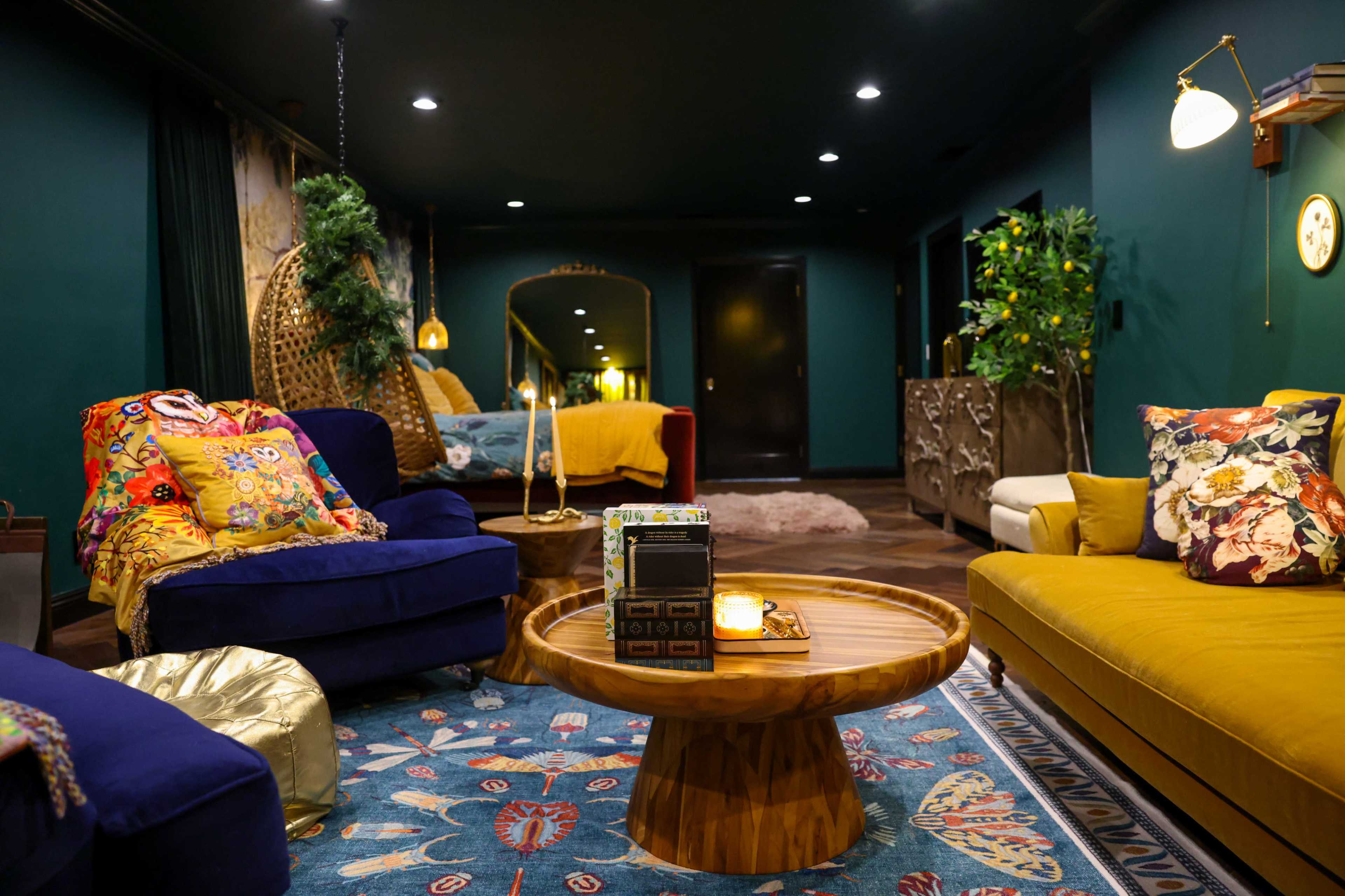 The image shows a cozy living space featuring a blue sofa, a yellow couch, a round wooden coffee table, and colorful decorative pillows, all set against dark green walls.