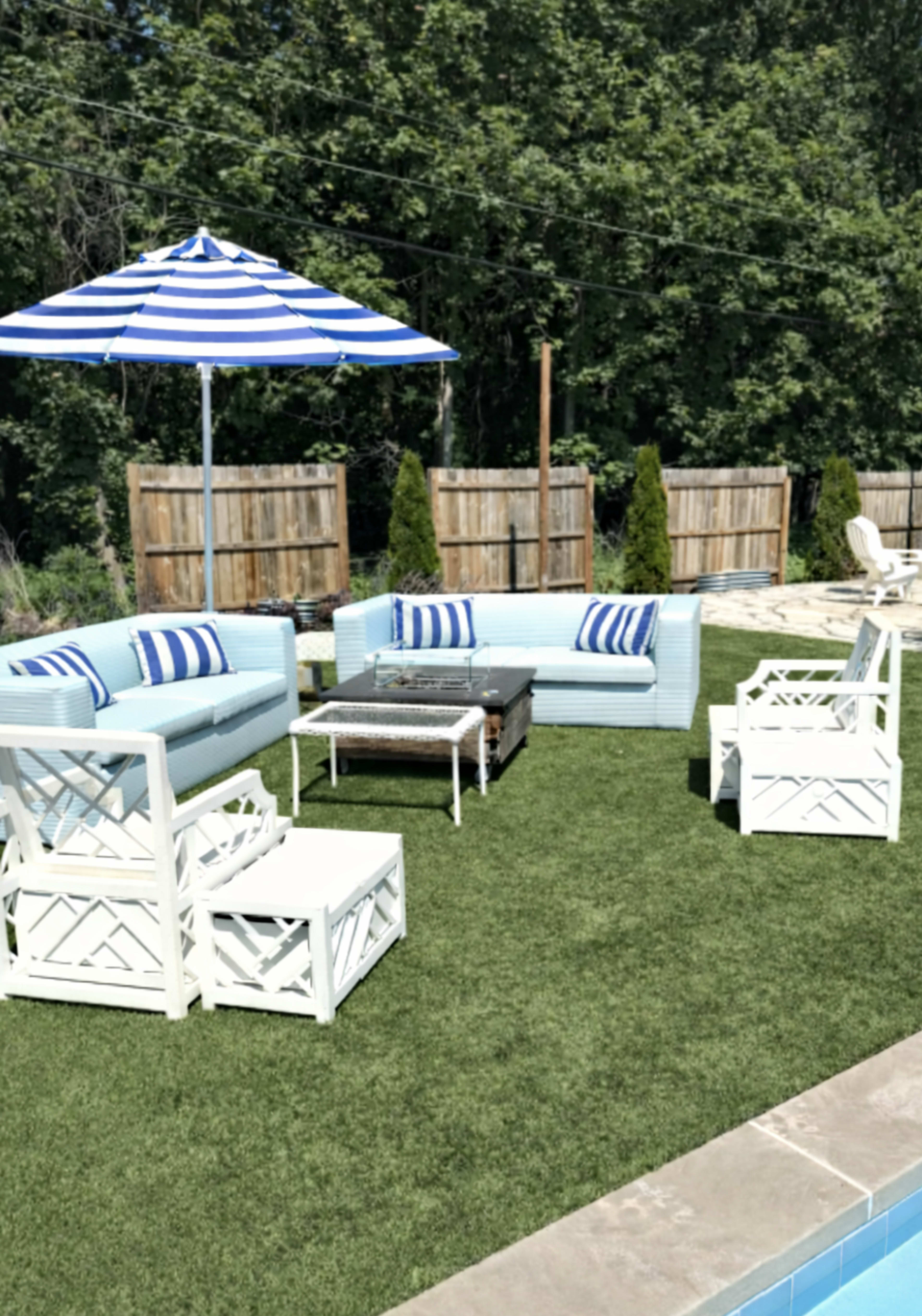 The image shows a backyard patio area with light blue outdoor sofas, a striped umbrella, a fire pit table, and white lounge chairs on synthetic grass.
