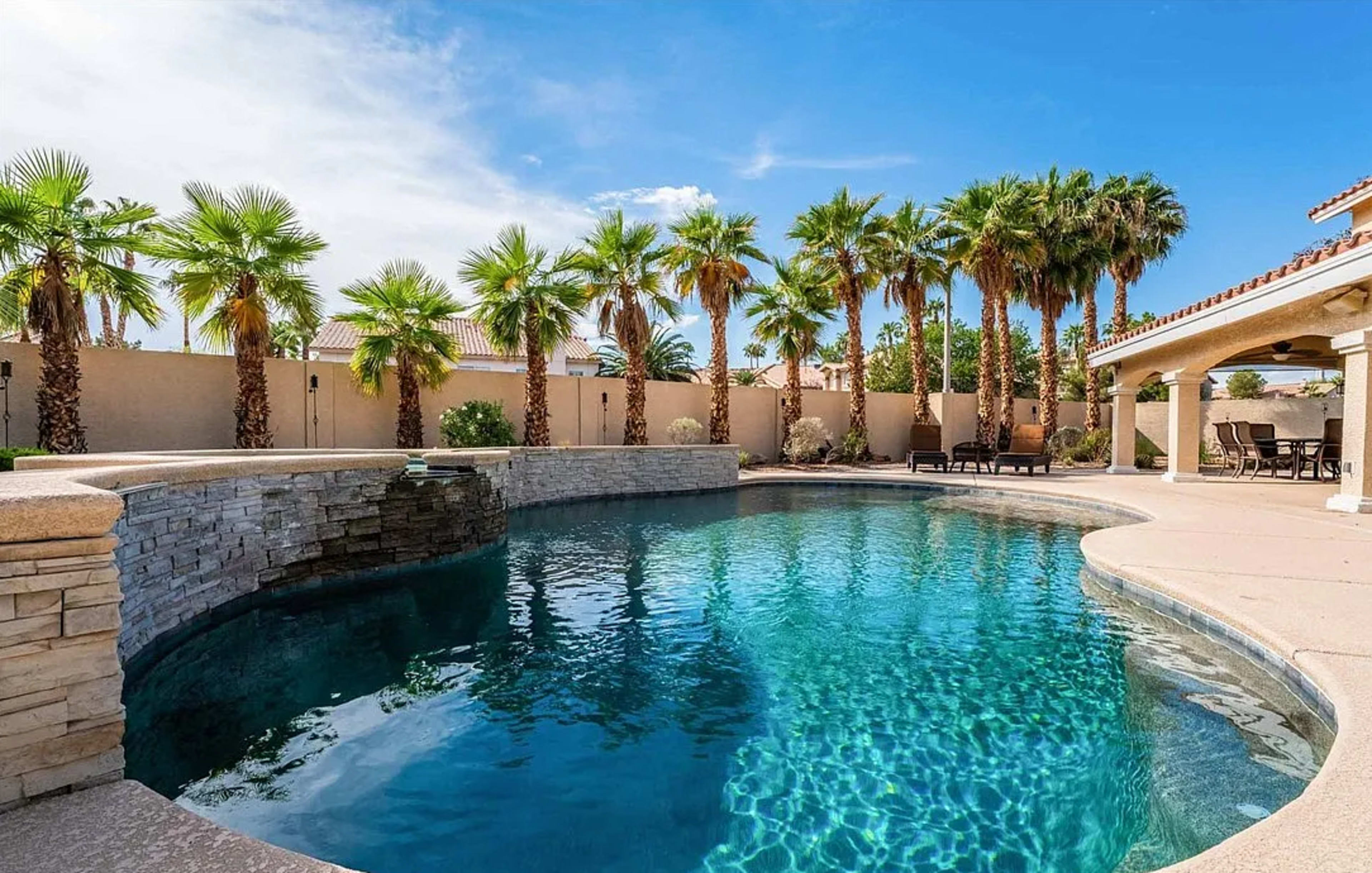 The image shows a swimming pool surrounded by palm trees and a patio area with lounge chairs under a clear blue sky.