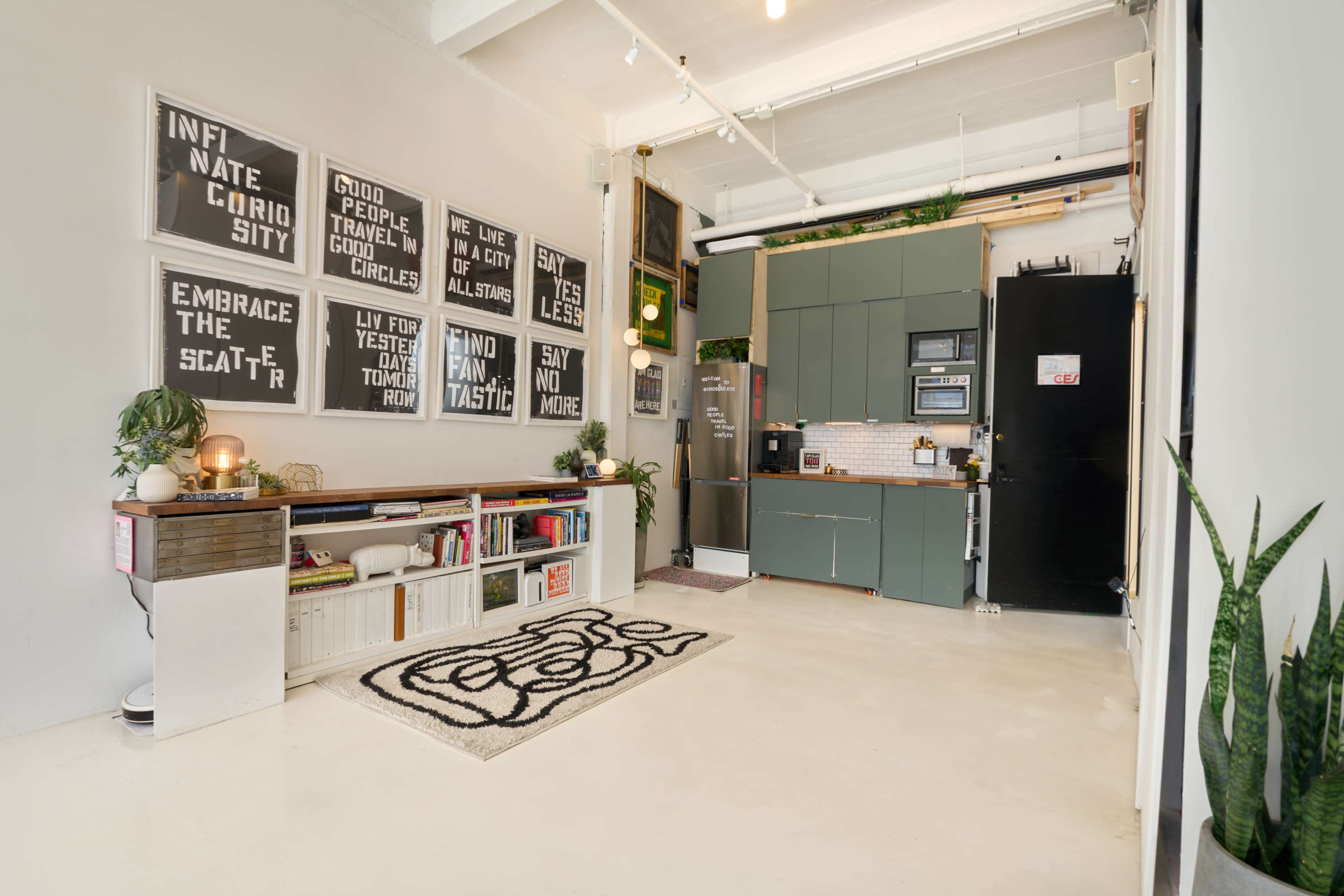 The image shows a modern, open living space featuring a kitchenette, wall art with motivational quotes, and a black and white patterned rug on a concrete floor.
