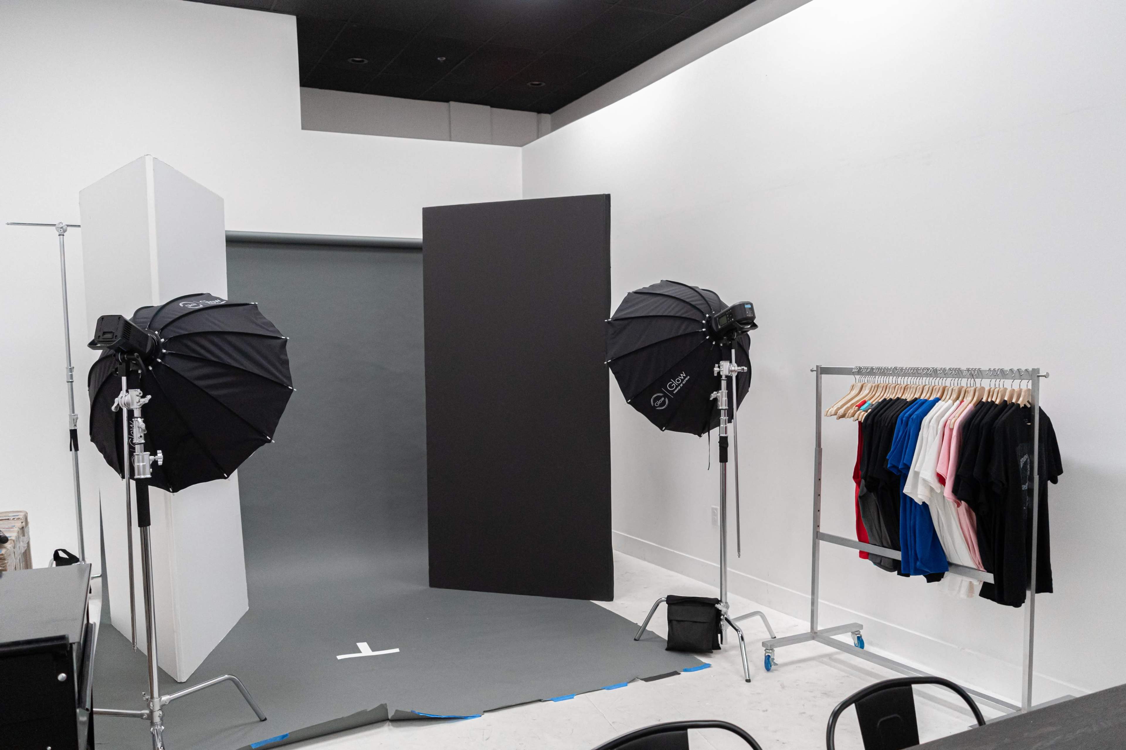 A photography studio setup with two lighting umbrellas, a backdrop, and a rack of assorted t-shirts in various colors.