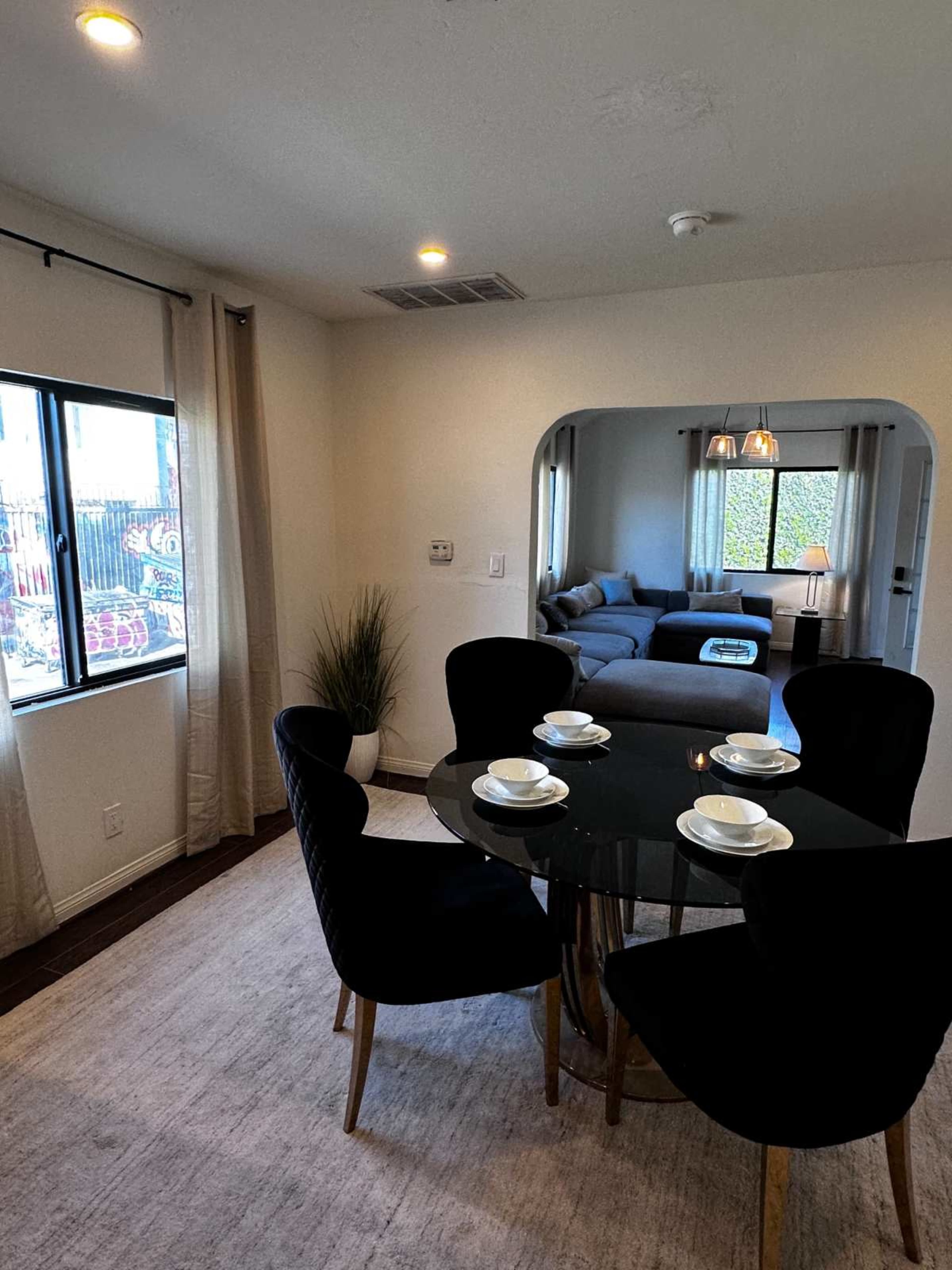 The image shows a dining area with a round black table set for four and a living room visible in the background.