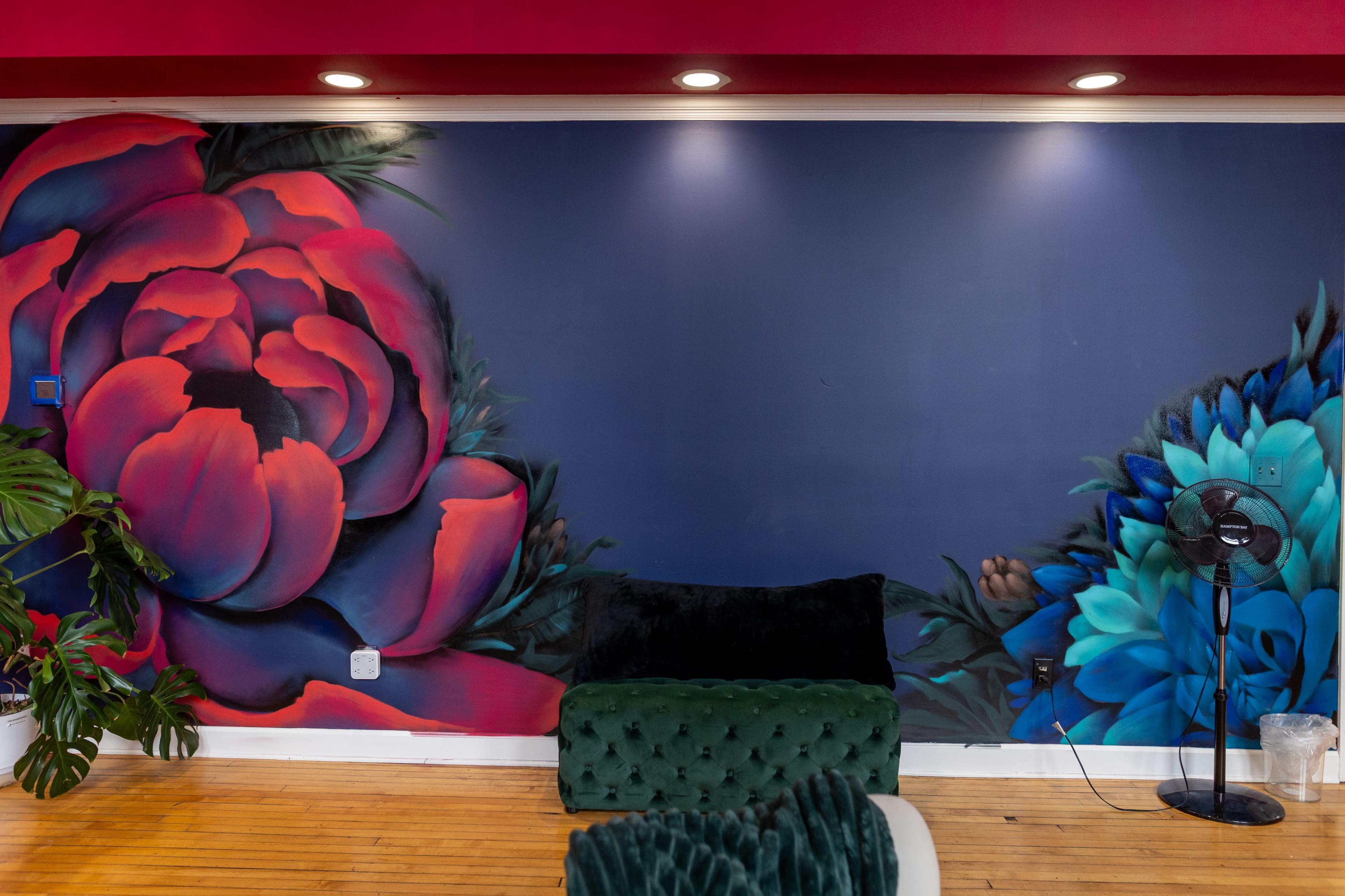 The image features a colorful mural of red and blue flowers on a dark blue wall, alongside a green velvet couch and a fan in a brightly lit room.