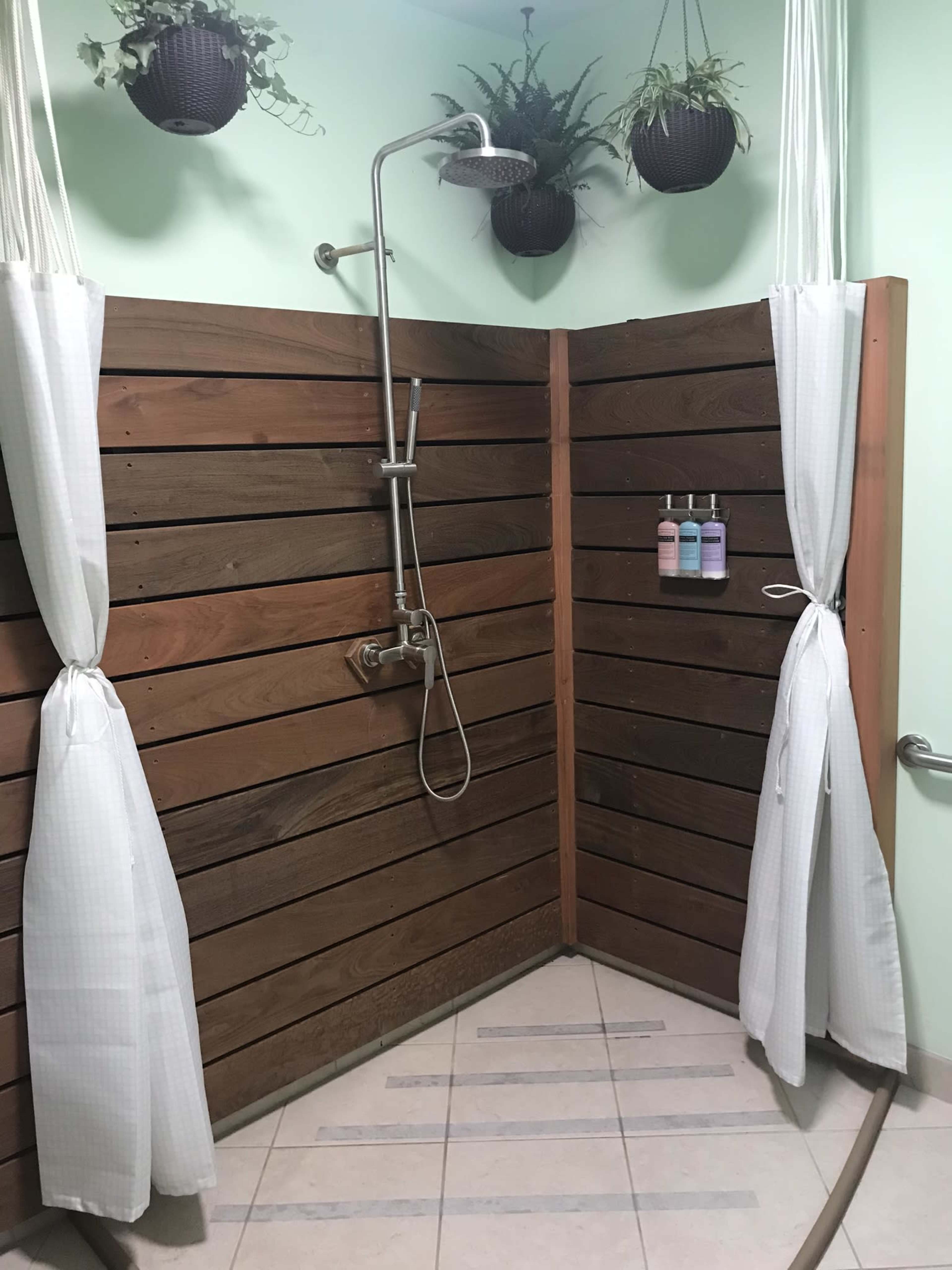 The image shows a modern shower area with wooden walls, a showerhead, and hanging plants above, along with three bottles of toiletries mounted on the wall.