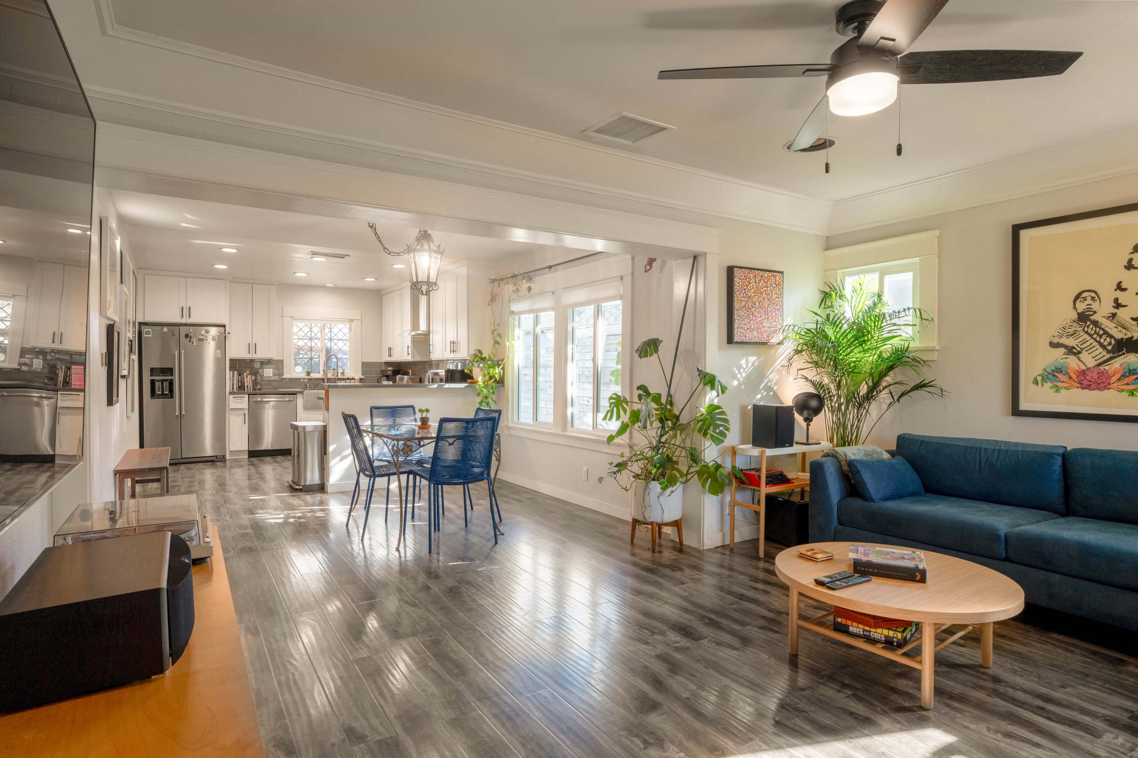 A modern living space features a blue sofa, wooden coffee table, and a dining area with metal chairs, surrounded by large windows and greenery.
