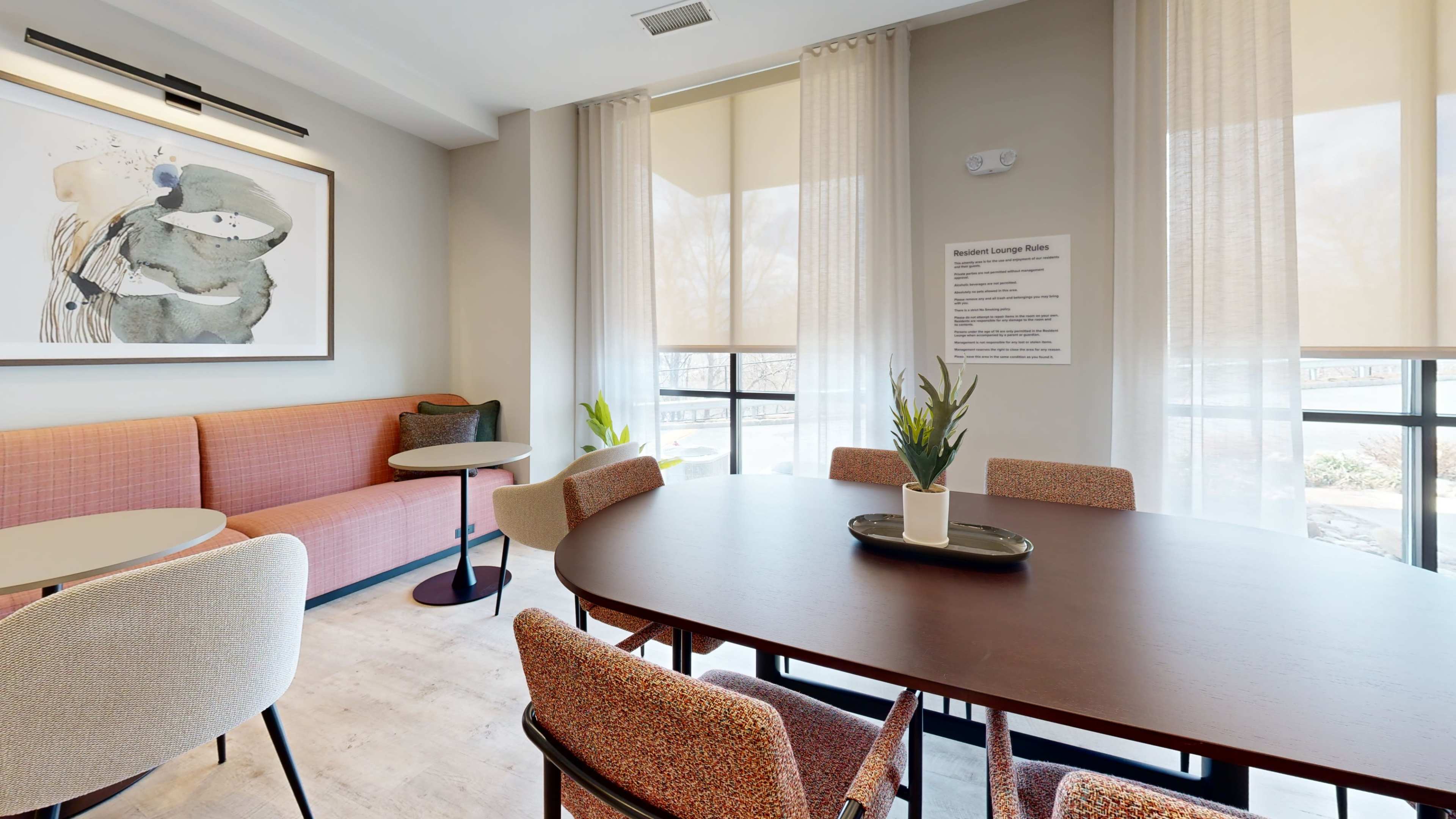 The image shows a modern lounge area with a round table surrounded by chairs and a sofa, featuring large windows with natural light and decorative plants.