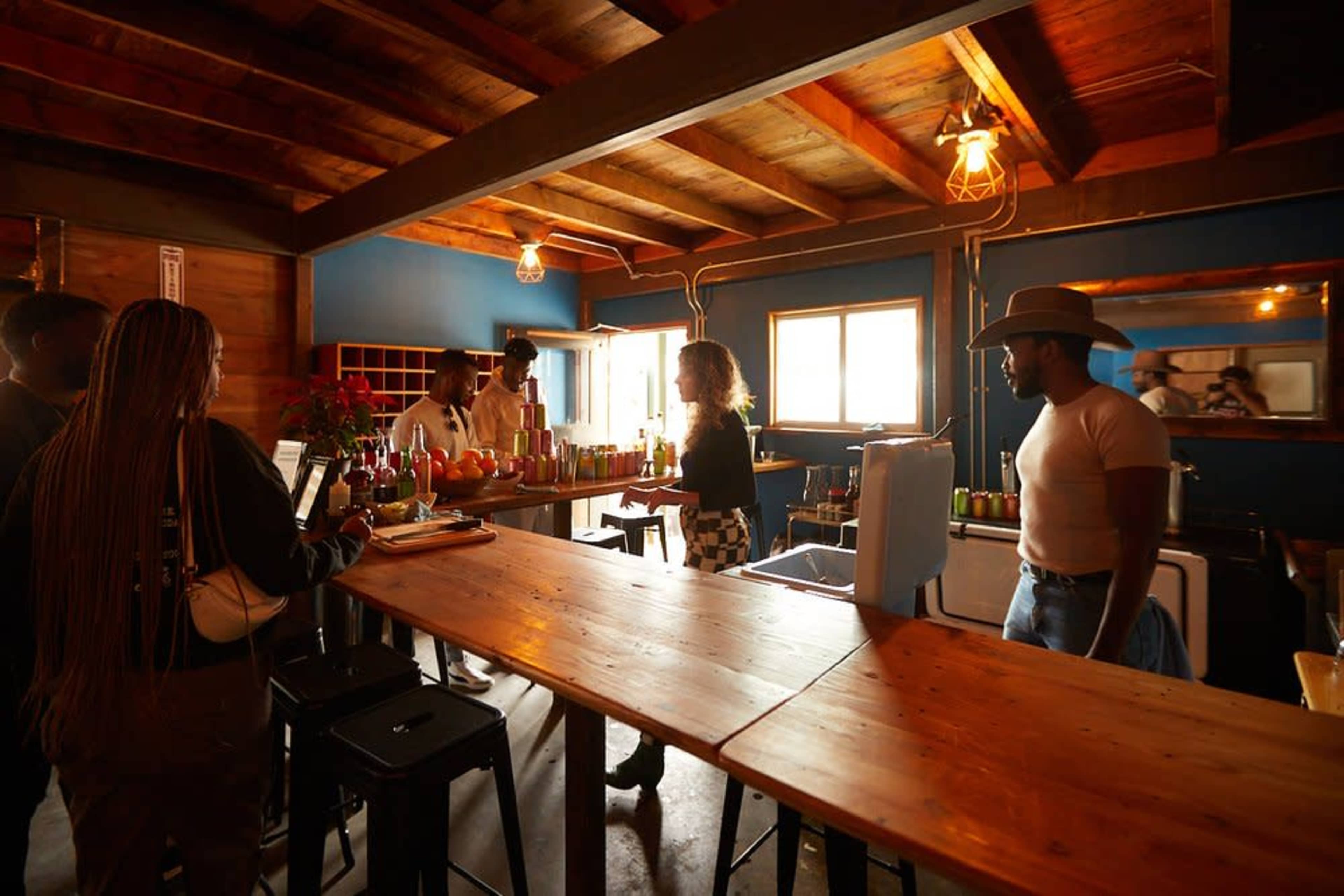 A group of people stands at a wooden bar in a cozy, dimly lit establishment with a blue wall and shelves filled with colorful drinks.