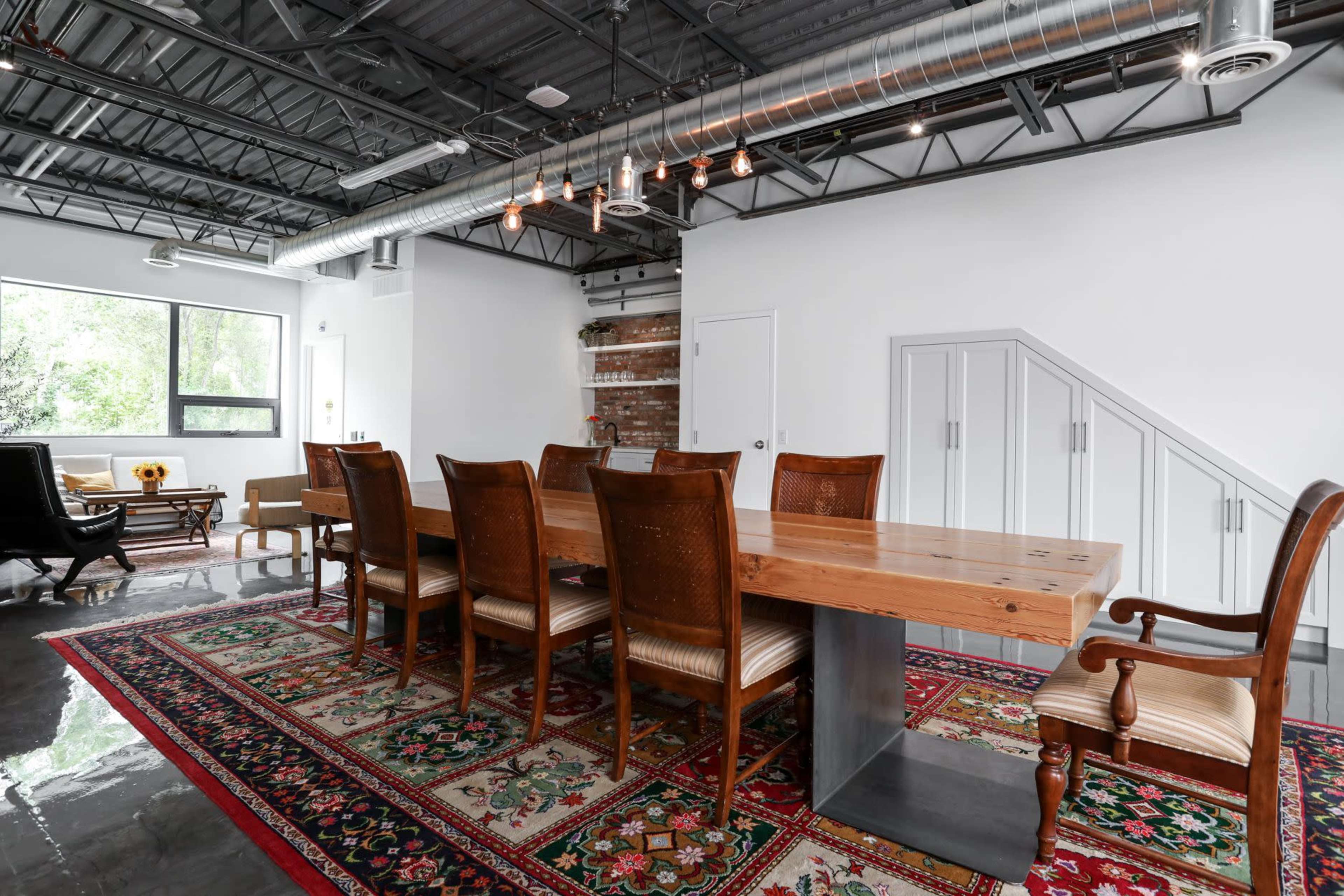 A long wooden dining table is surrounded by several chairs on a patterned rug in a brightly lit room with exposed pipes and a staircase.